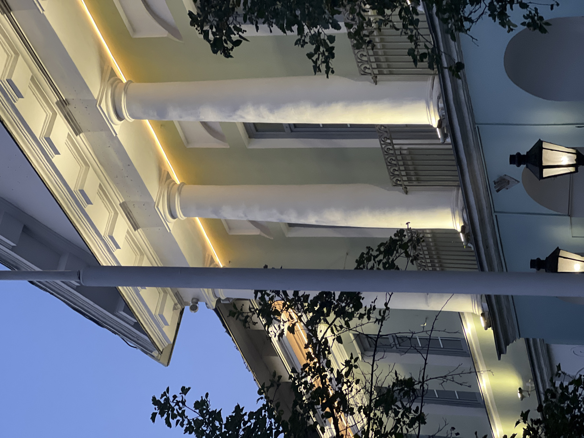 The image captures a nighttime view of a building facade in Tver, Russia. The building exhibits classical architectural elements, featuring large white Corinthian columns that support an upper story. A warm yellow light runs along the top of the facade, illuminating the decorative cornices and highlighting the columns. Several windows with grey frames are visible behind the columns. A balcony with an ornate metal railing runs along the facade, partially obscured by dark green foliage from trees. The lower section of the building is painted a light blue and features arched openings, with a vintage-style lamppost casting a warm glow in the foreground. The sky above is a deep twilight blue.