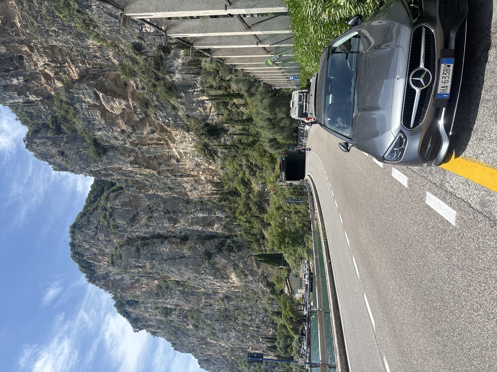 A dark gray Mercedes-Benz C-Class sedan is parked on the shoulder of a two-lane asphalt road that curves around a steep, tree-covered mountain. The car is angled towards the camera, showing its front grille with the Mercedes-Benz logo and a license plate that reads "HA-633BW". The road is marked with white lane lines and a yellow line along the edge where the car is parked. To the left of the road, a body of water, likely a lake, is visible with several boats docked. The mountain face is rocky and rugged, with patches of green vegetation and trees growing on its slopes. In the distance, a tunnel entrance is visible in the side of the mountain, with a motorcycle and a camper van approaching it. The sky is blue with scattered white clouds. The scene appears to be in a mountainous, coastal region. The location is identified as being in the city of Tignale, Italy.