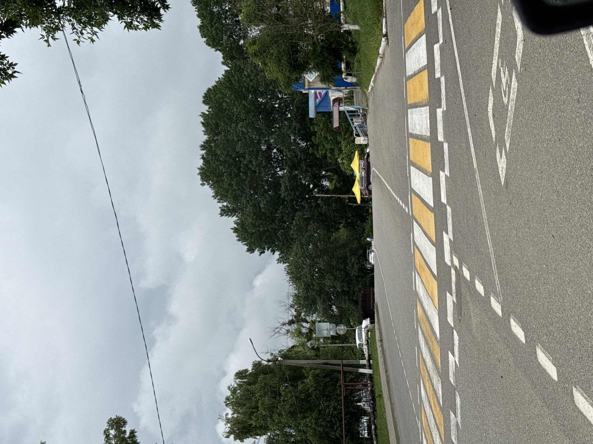 The image shows a cloudy day scene on a road in Tatarstan, Russia, likely near a residential or commercial area. A crosswalk with alternating yellow and white stripes is prominently featured in the foreground. To the left of the crosswalk, a sidewalk runs alongside a grassy area, with a fence and some trees visible. Beyond the sidewalk, a white car is parked.
In the middle ground, a road curves to the right. A street lamp stands on the left side of the road. Several trees of varying heights and densities form a backdrop. Two yellow awnings are set up near a blue building with a sign displaying Russian lettering. A dark-colored car is parked in front of the awnings. Further down the road, another car is visible, along with a person standing on the sidewalk. The road surface is asphalt, and there are white dashed lines indicating lanes.