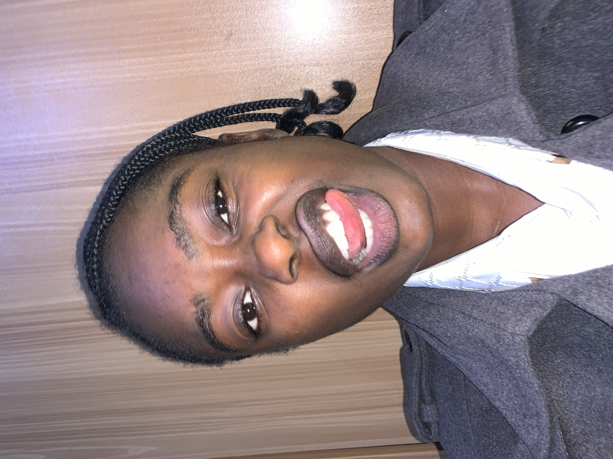 A young Black woman with braided hair styled in pigtails is shown in a close-up, selfie-style portrait. She is wearing a dark gray coat over a white collared shirt. Her mouth is open with her tongue sticking out, and her eyes are looking towards the camera. The background appears to be a light brown wooden wall. The image is lit from the front, casting a slight shadow on the left side of her face. The context of this image, including its location in Gura Top, Nigeria, is not evident from the visual information alone.