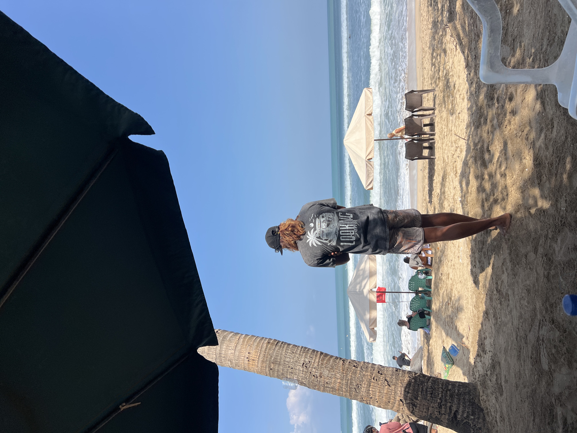 The image depicts a beach scene in Kuta, Indonesia, on a bright, sunny day. In the foreground, a dark green umbrella partially obscures the top of a tall, rough-barked palm tree. A man with long, reddish-brown hair and wearing a black baseball cap stands with his back to the camera, arms crossed. He is wearing a dark grey t-shirt with a graphic design that includes the text "LOOKING GOOD". He also wears patterned shorts.
Beyond the man, the sandy beach is lined with beach chairs and umbrellas. Two beige umbrellas are visible, one with a red flag. Several people are scattered on the beach, some sitting in chairs under umbrellas and others walking near the water. The ocean has moderate waves, and several surfers can be seen in the distance. The sky is clear blue. The sand is light brown, and shadows from the foreground umbrella and palm tree are cast across it. In the lower right corner, a white plastic beach chair is partially visible.