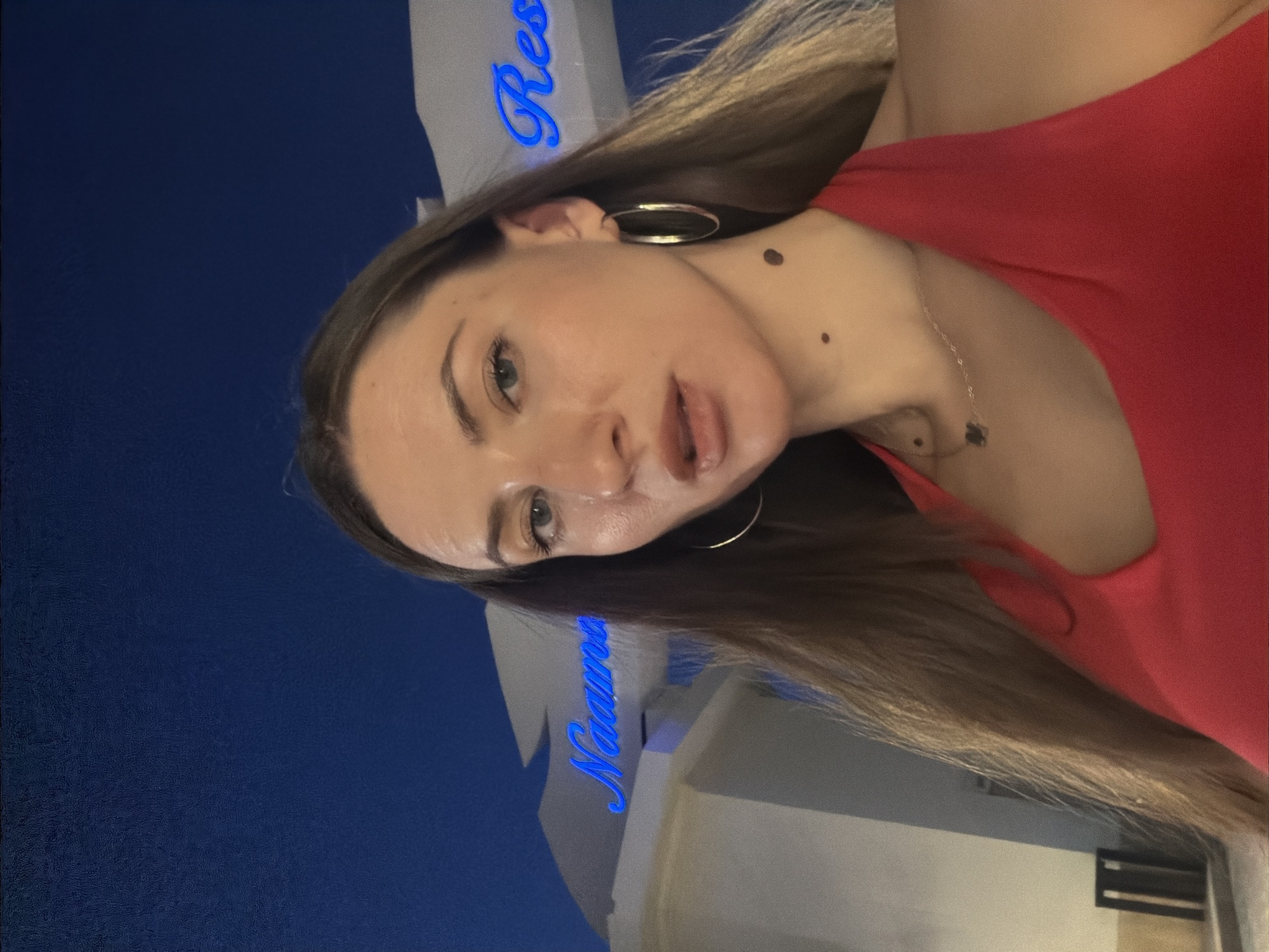 The image is a selfie taken at night outside a resort in Sharm El Sheikh, Egypt. A woman with long brown hair and fair skin is looking at the camera. She is wearing a red halter top and large hoop earrings. Behind her, illuminated by blue neon lights, are signs that read "Naama" and "Res". The background appears to be the facade of a building, possibly an entrance to the resort. The sky is a deep blue, indicating it is nighttime.