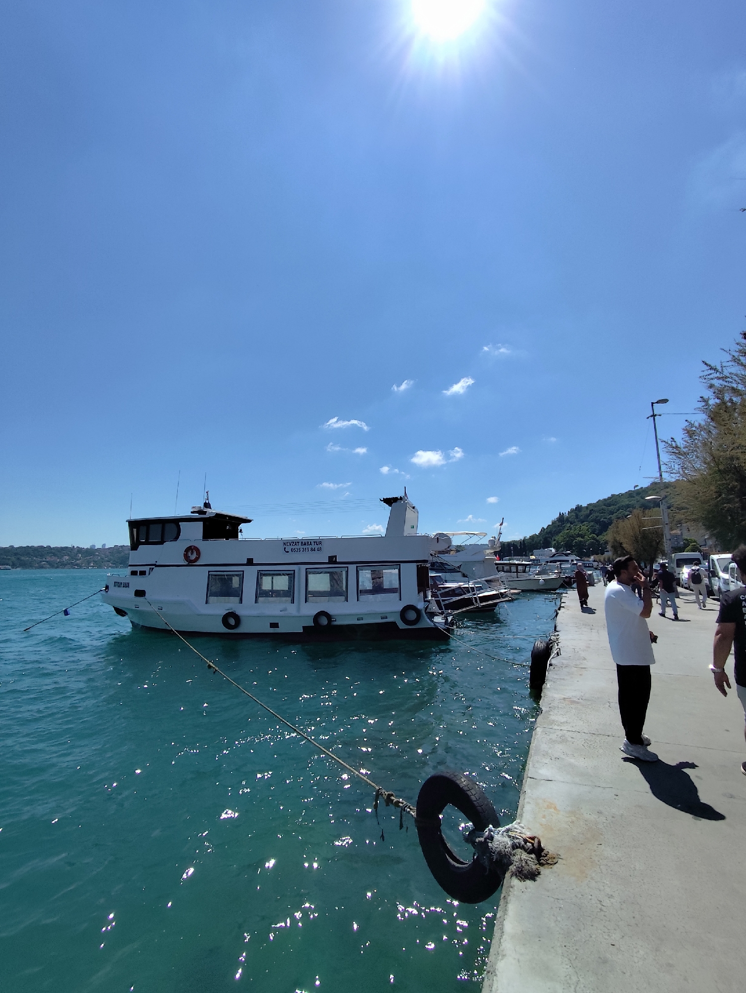 A wide shot captures a sunny day at a harbor. A white passenger boat, the "NEVZAT BABA TUR" with a phone number "0535 319 84 48" visible, is docked on the left side of the frame, its bow facing left and slightly away from the viewer. The boat has several windows and a life preserver attached to its side. The water is a vibrant turquoise, reflecting the bright sunlight. A concrete pier runs along the right side of the frame, with several people standing and walking on it. One man in a white shirt and black pants is facing away from the viewer, looking out at the water. Another person in a black shirt is partially visible on the far right. Mooring lines and tires are used to secure the boat to the pier. In the background, a lush green hillside slopes down towards the water, dotted with trees and some buildings. The sky is a clear, bright blue with a few wispy clouds and the sun prominently visible in the upper left corner, casting a strong glare. Several other boats are moored in the background, and power lines stretch across the sky.