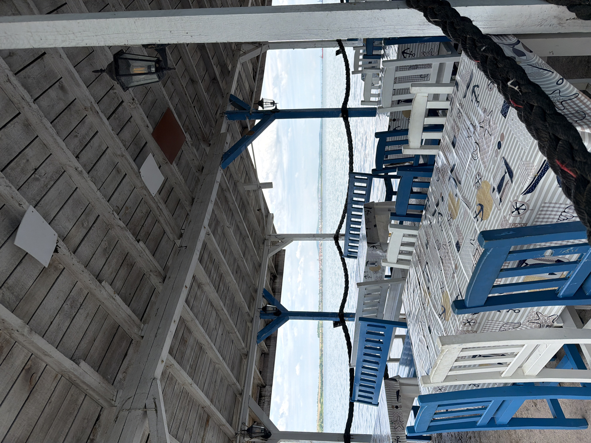The image shows a view from under a wooden structure, possibly a covered terrace or restaurant, looking out over a body of water. The structure has white and light-colored wooden supports and a roof. There are hanging lamps attached to the wooden beams. Below, there are tables covered with tablecloths featuring a nautical theme, such as anchors, starfish, and seashells. The tables are surrounded by blue and white wooden chairs. A thick, dark rope is draped across the scene, adding a nautical touch. In the distance, across the water, there's a visible coastline with some greenery and buildings. Based on the style of the restaurant, the color scheme, and the general layout, this scene could be located in the city of Burgas, Bulgaria, along the coast of the Black Sea. There are no people visible in the shot.