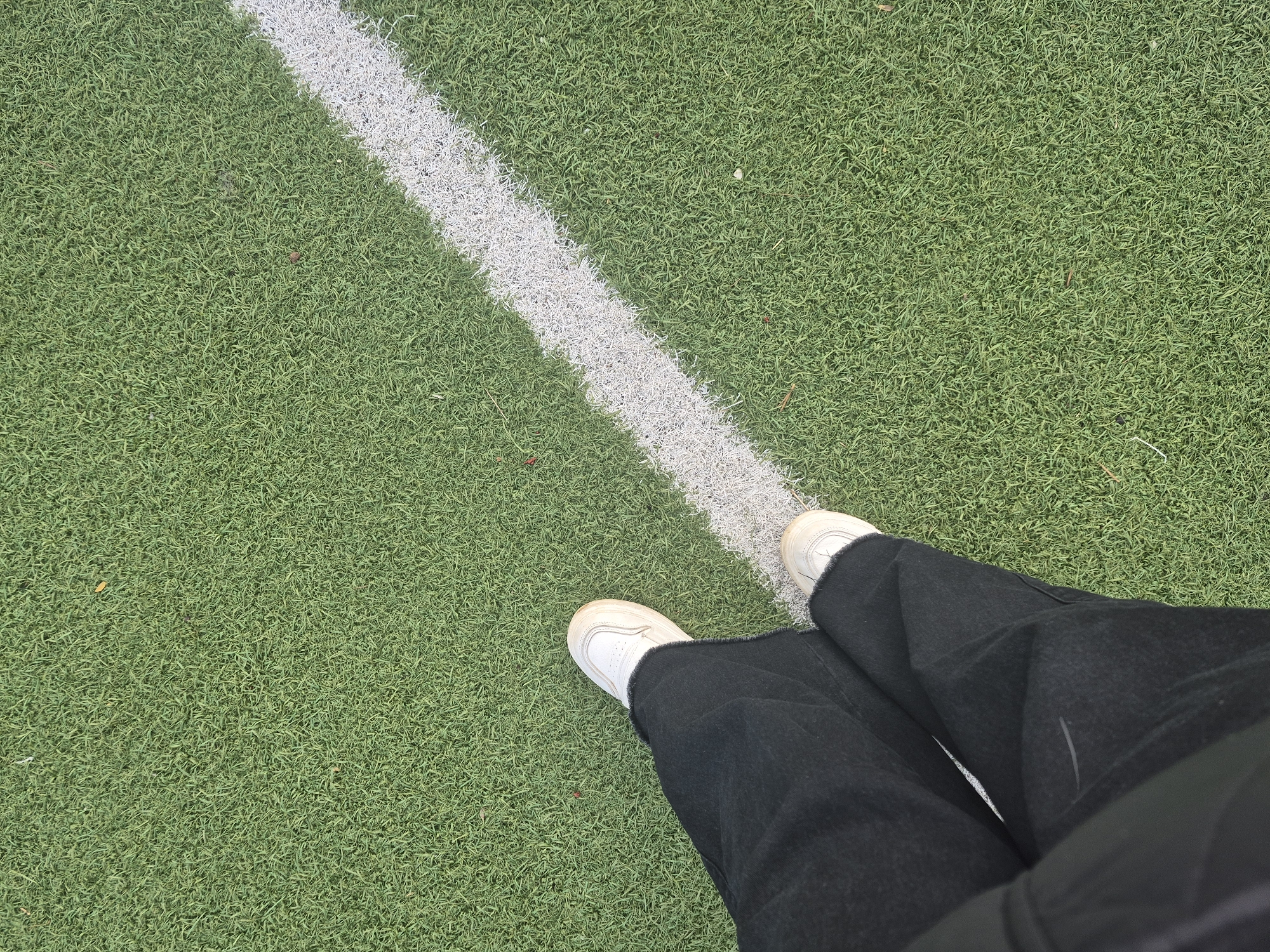 The image presents a top-down view of a person's lower body on an artificial green turf field in Chaikovsky, Russia. The individual is wearing loose-fitting, black wide-legged pants and clean white athletic sneakers. Their feet are positioned very close to, or slightly over, a distinct white boundary line that cuts diagonally across the green field from the upper right to the lower left.
The field itself is covered in short-pile artificial green grass, which appears uniform in texture. The white line, contrasting sharply with the green, exhibits a slightly textured or frayed appearance, typical of painted lines on artificial turf. The lighting suggests it is daytime, casting soft, indistinct shadows. No specific activity or event is taking place other than the person standing on the field. No visible text is present in the media file.