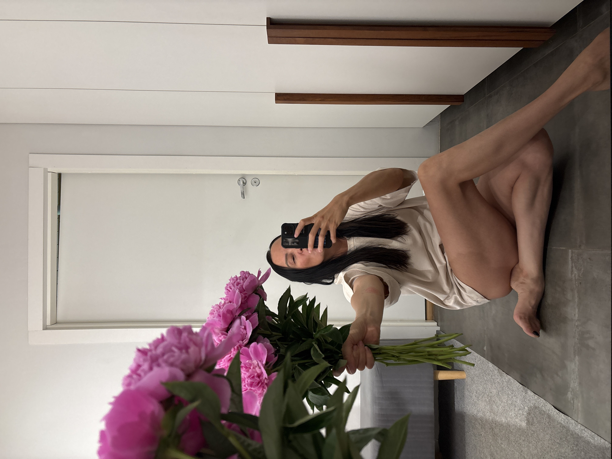 A person is kneeling on a gray tile floor in an indoor location. They are holding a bouquet of pink peonies in their left hand and taking a photograph of themself with a black smartphone in their right hand. The person has dark hair and is wearing a light-colored, loose-fitting top. Behind them is a white door with a silver handle and a white wall with dark wood trim near the ceiling. In the background, there's a gray upholstered bench. The location is Chaikovsky, Russia.