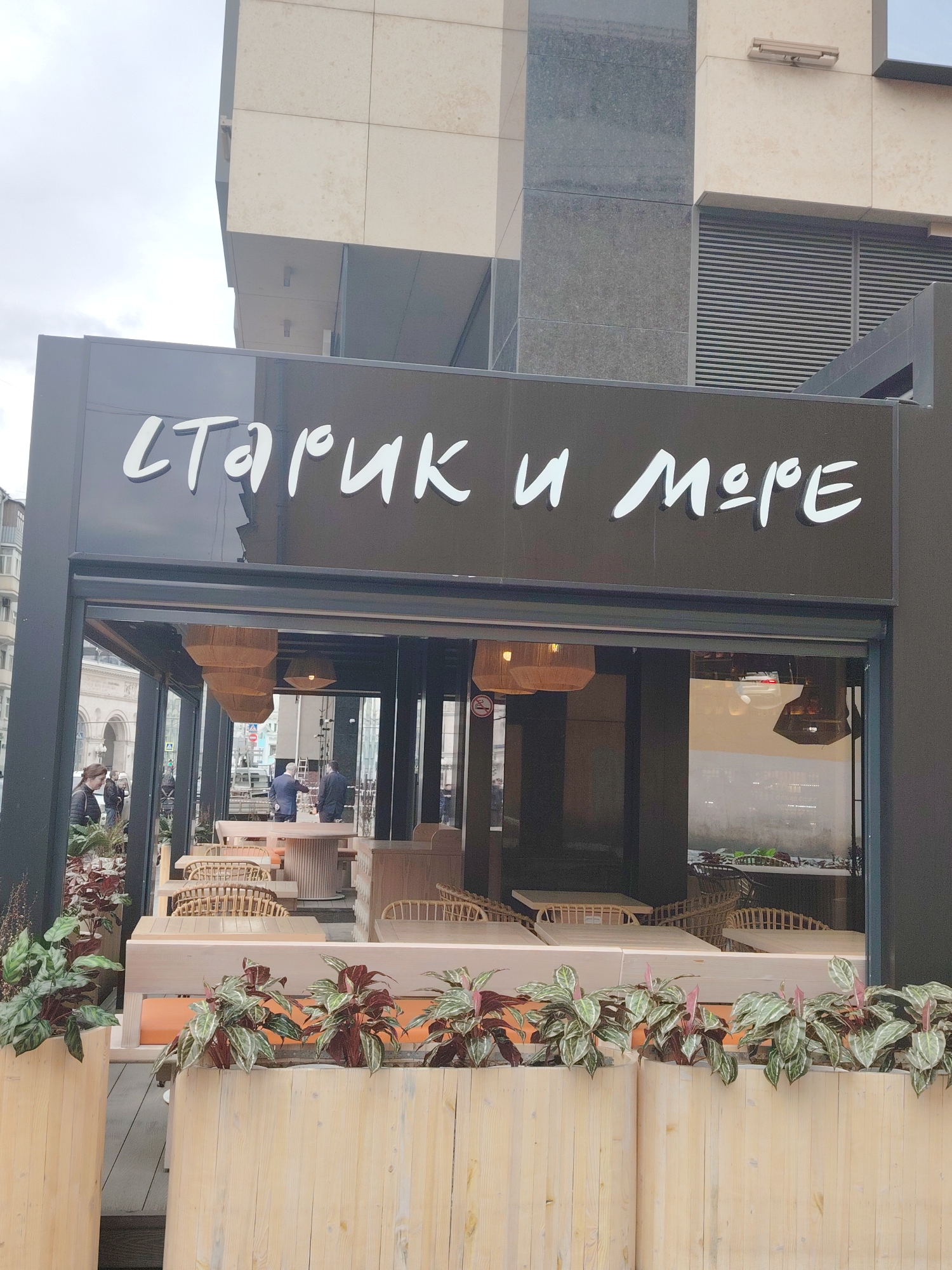 The image captures the exterior of a modern restaurant named "СТАРИК И МОРЕ" (Starik i More, meaning "The Old Man and the Sea" in Russian), located in Moscow, Russia. The restaurant's sign, displaying the name in white, stylized Cyrillic letters, is prominent against a dark facade.
Below the sign, a glass-enclosed dining area is visible, extending into an outdoor patio with additional seating. The patio features multiple light-colored wooden planters filled with green and maroon-leafed plants, creating a natural boundary. Within the patio area, light-toned wooden tables and benches are paired with wicker-style chairs, suggesting a contemporary and relaxed dining atmosphere.
Through the large glass windows, several people are visible inside the restaurant. One individual appears to be standing near a table, possibly a staff member, while others are less distinct in the background. On the street outside the restaurant, near the left edge of the frame, a person is walking past.
The restaurant is housed within a larger, modern building characterized by sections of light-colored and dark-grey paneling, and a large vent or grille is visible on the upper part of the building's facade. The sky above is overcast, indicating a daytime scene. The overall impression is that of an inviting urban eatery with a pleasant outdoor space.
