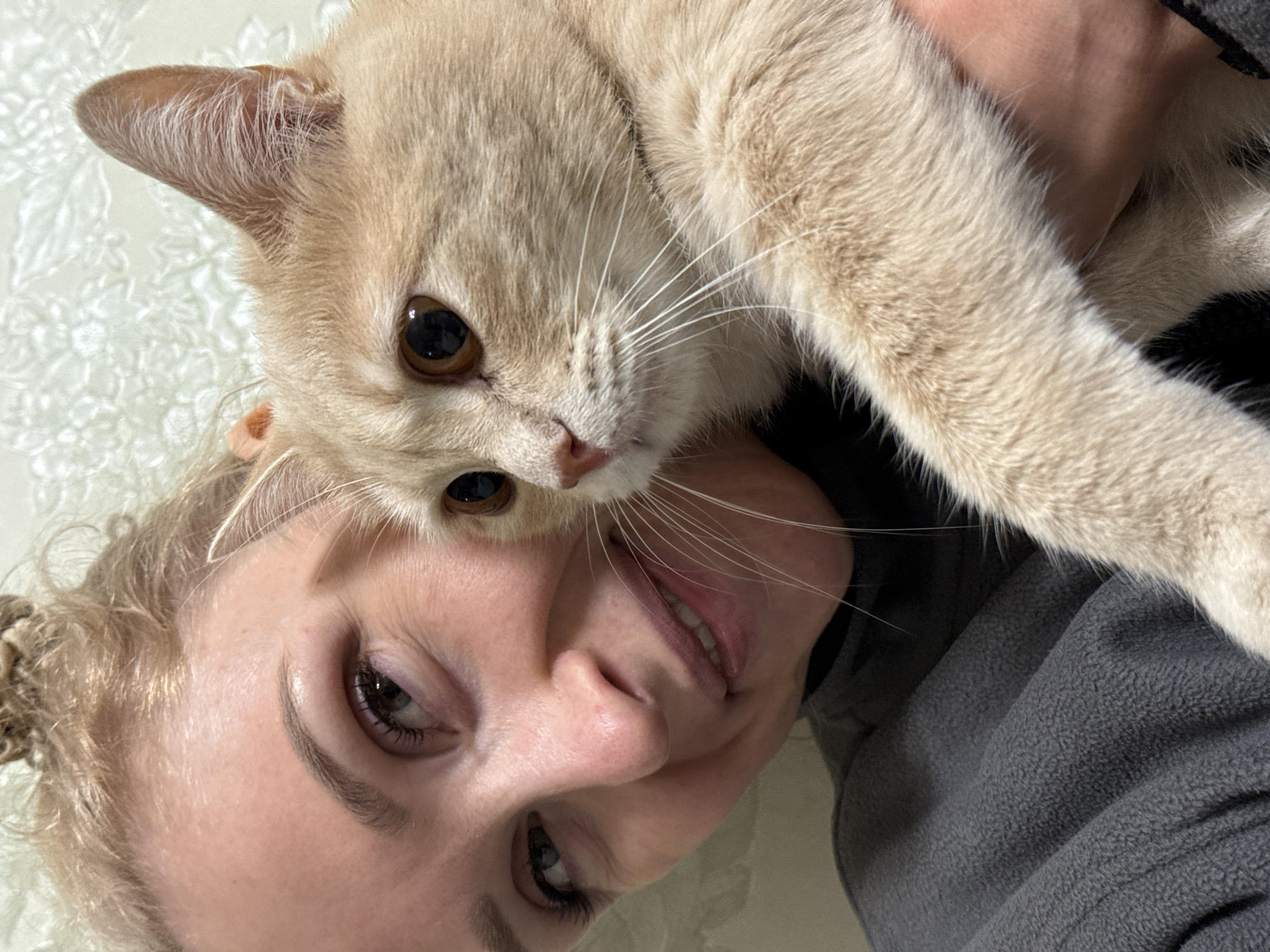 A selfie of a woman with a cat. The cat is light brown with darker brown eyes. The woman has blonde hair pulled back and brown eyes. She is wearing a gray fleece jacket. The background is a light green patterned surface. The image was likely taken indoors and is rotated counter-clockwise. There is no indication of the city of Perm, Russia.