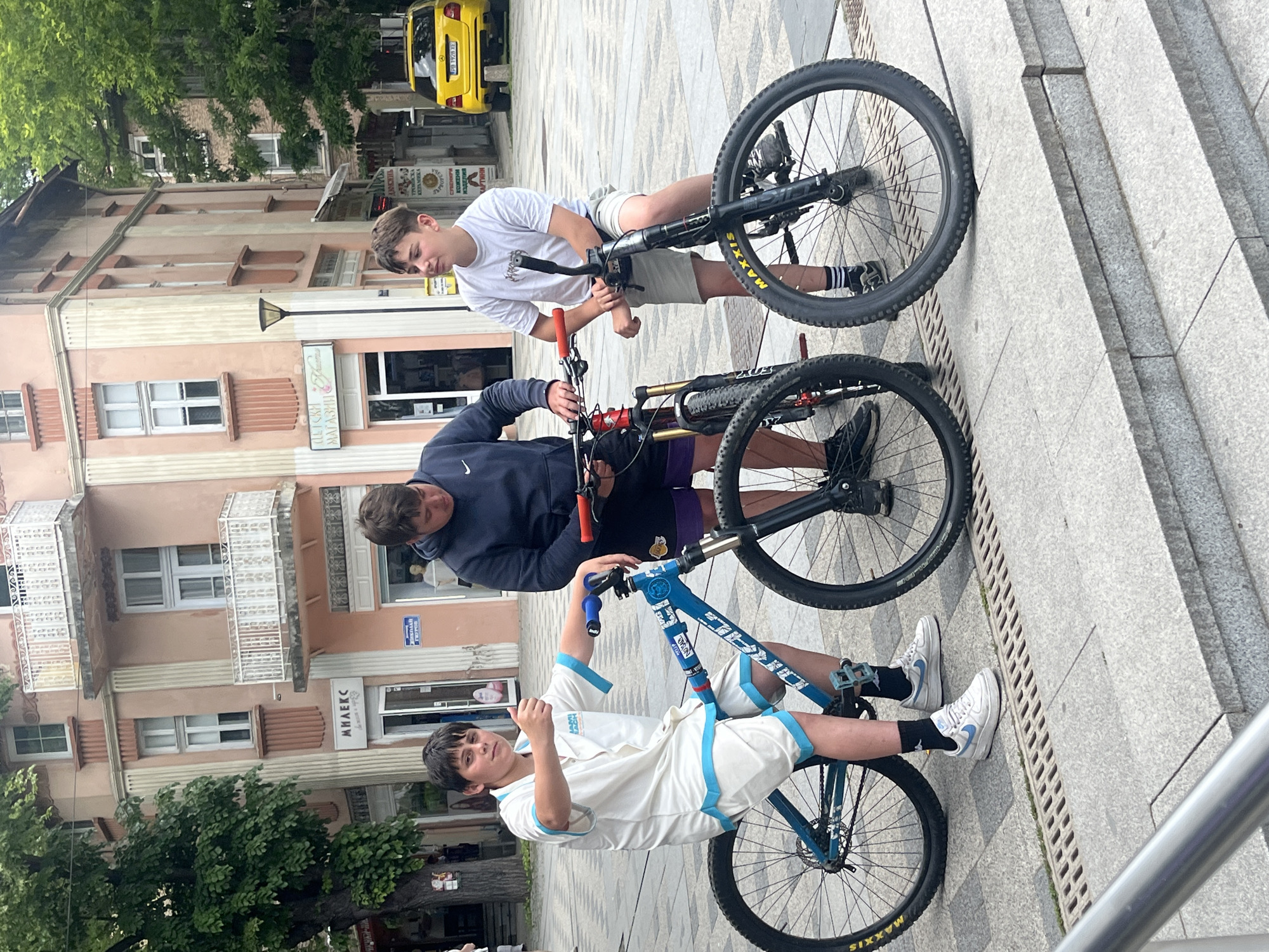 The image depicts three young males with their bicycles in an outdoor urban setting within the city of Unknown, Unknown.
In the foreground, grey granite-like paving stones feature two parallel drainage grates. A series of three grey stone steps ascend from the left.
From left to right:
The first male, of young age, is seated on a blue "DRAG" brand hardtail bicycle. He wears a white button-down shirt with light blue trim on the collar and sleeves, matching white shorts, white Nike Air Force 1 sneakers with light blue accents, and black socks with a white logo. He has dark hair and is looking towards the viewer, holding his left thumb up in a 'thumbs-up' gesture. The bicycle has "MAXXIS" tires with yellow lettering and blue handlebars/grips.
The second male stands centrally, positioned between his red full-suspension mountain bike and the third male's black hardtail bicycle. He wears a dark blue hooded sweatshirt with a white Nike swoosh, purple athletic shorts displaying a yellow "LAKERS" logo, black sneakers, and black socks. His attention is directed downwards towards the front wheel or suspension of his red bicycle.
The third male stands to the right, next to his black hardtail bicycle, which also features "MAXXIS" tires with yellow lettering and red handlebars/grips. He wears a white t-shirt with a dark graphic, light grey shorts, black sneakers, and black socks with white stripes. He has dark hair and is looking towards the right side of the frame, away from the viewer.
The background features a street with various multi-story buildings, primarily in shades of peach and light pink. These buildings have white-framed windows and decorative white balconies with railings. Several ground-floor storefronts are visible. On the left building, a sign reads "МИЛЕКС" (MILEX) in Cyrillic. On the building immediately behind the middle and right males, signs in Cyrillic script include "ЦВЕТНИ МАГАЗИНИ" (Flower Shops) with a stylized graphic, and below that, "ТРОЯНСКА КЕРАМИКА" (Troyan Ceramics), "СУВЕНИРИ" (Souvenirs), "КОЖЕНИ ИЗДЕЛИЯ" (Leather Goods), and "КАРТИНИ" (Paintings). Another sign reads "БУЛГАРИЯ ГОТОВ" (Bulgaria Ready). A black street lamp with a modern design is situated between the central buildings. Further back, a bright yellow Mercedes-Benz SUV is parked, displaying a European-style license plate with the registration "PB 1928 XE". Lush green trees are visible on both sides of the street. The lighting suggests an overcast or diffused daylight environment.