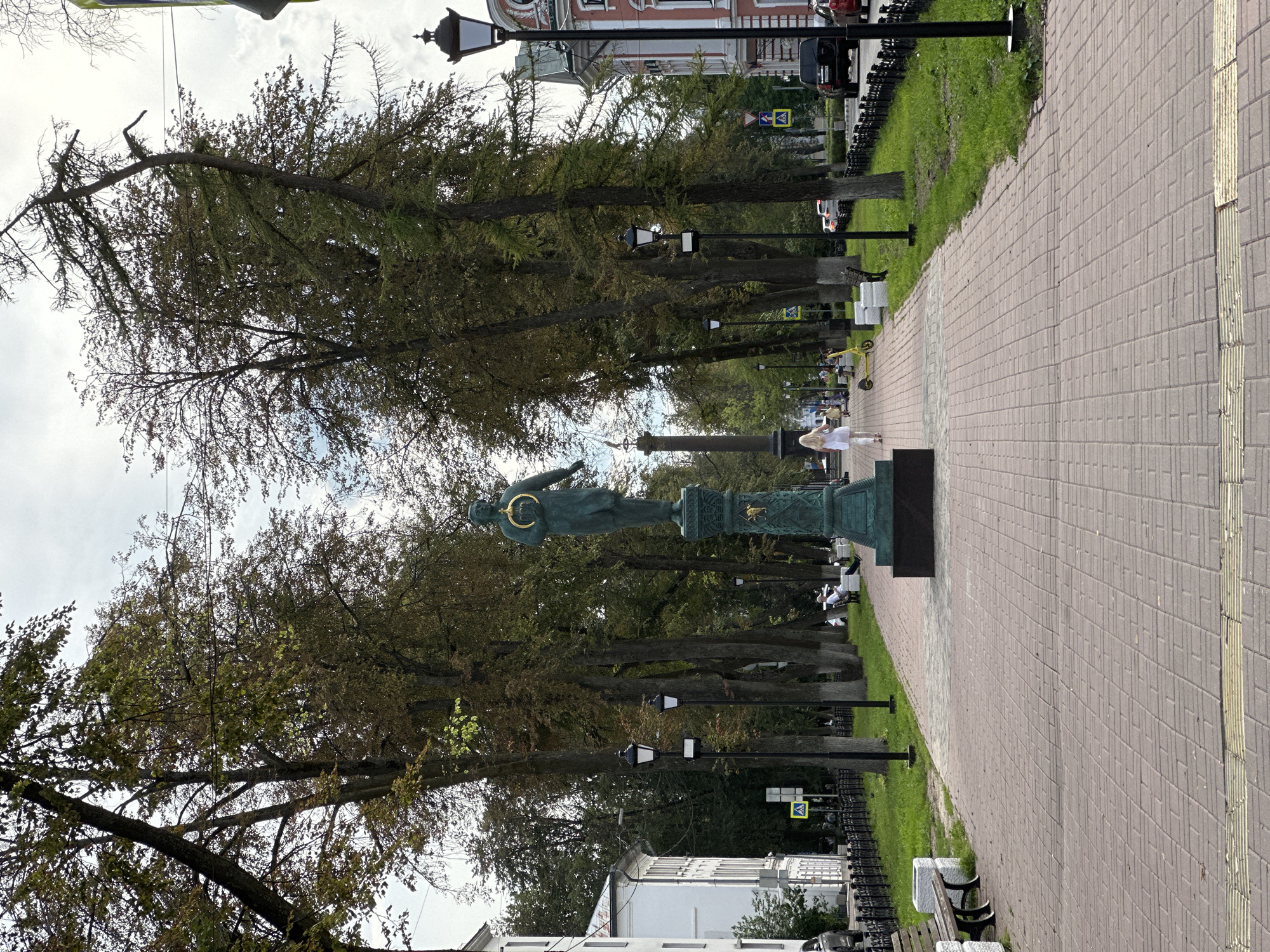 The image depicts a street scene in the city of Zhukovo, Russia. In the foreground, a brick-paved walkway runs through a tree-lined park. A statue of a person holding a golden ring stands prominently in the center of the walkway. Several lampposts are visible along the path, and benches are placed on the grassy area to the left. In the background, a building with white walls and windows can be seen on the left, and a building with pink and white facade and windows is visible on the right. Cars are parked and driving on the street beyond the park. A person in a white top and light-colored pants is walking away from the camera on the walkway. Other individuals are seated on benches or walking further down the path. A yellow scooter is parked on the grass. The sky is overcast with clouds.