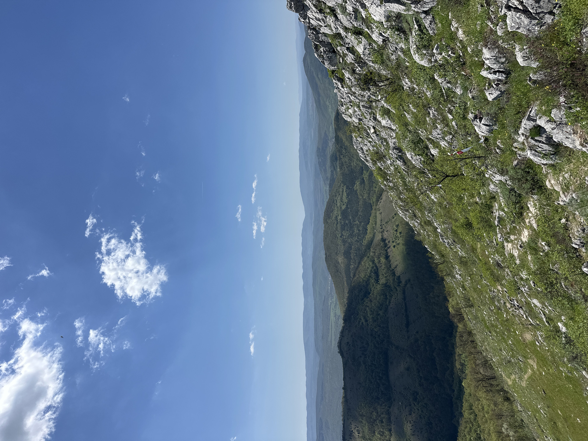 The image depicts a landscape from a high vantage point, likely a mountain in the Bilka area of Bulgaria. The sky occupies the left portion of the frame, a clear blue with scattered cumulus clouds. The right side features a steep, rocky cliff face covered in green vegetation, with exposed grey rock formations. In the distance, layers of mountains fade into a hazy blue, with deep green valleys visible between them. There are no people visible in the scene. A small, red and white object is visible on one of the branches. The image is taken during daylight hours in clear weather.