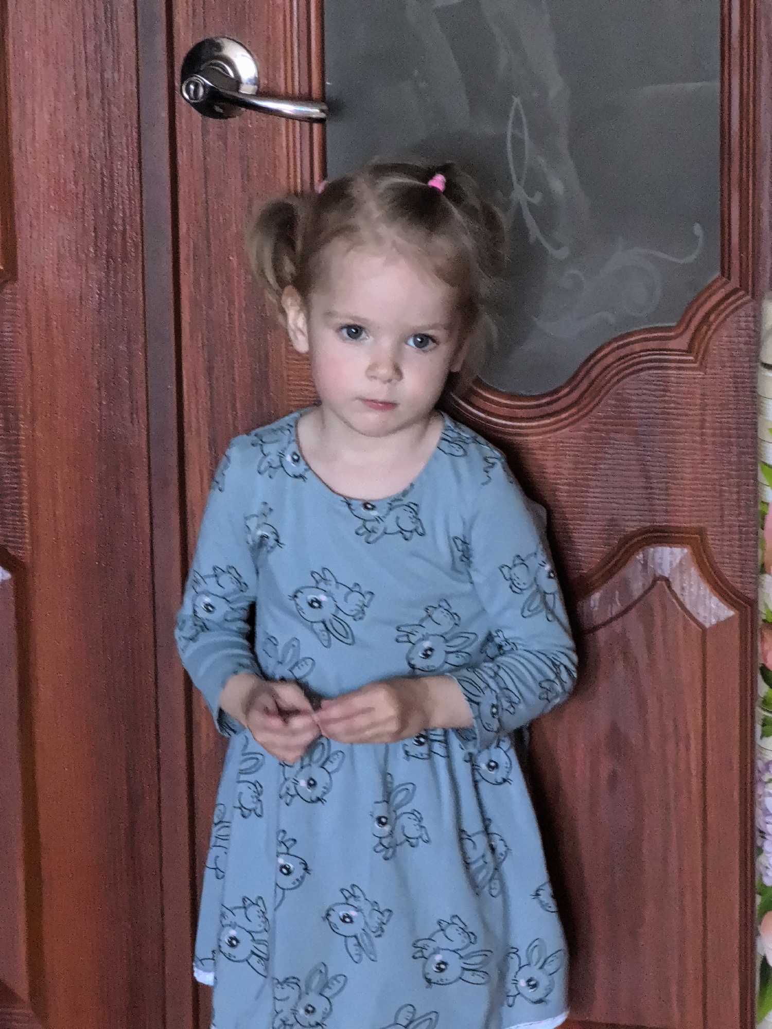 A young girl stands facing forward in front of a wooden door. The girl has blonde hair styled in two pigtails, a light blue dress with a pattern of bunnies, and blue eyes. Her hands are clasped in front of her. The door is dark brown with a silver doorknob on the left side. To the right of the door, there is a dark pane of glass with a white decorative swirl pattern. The lighting is natural and appears to be from the front. The overall scene is indoors.