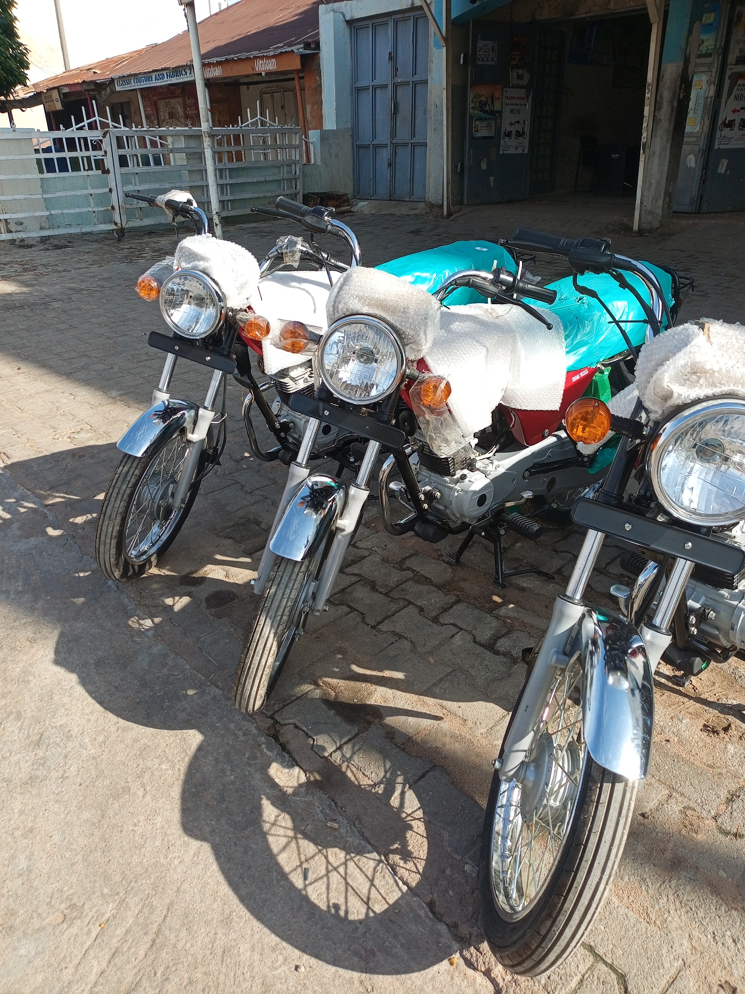 In Arawa, Nigeria, on a paved street, three motorcycles are parked outside a building. The motorcycles are new and appear to be for sale, with some parts covered in protective material. Two of the motorcycles are visible in the foreground, facing towards the viewer, while the third is partially obscured behind them. The building in the background has a corrugated iron roof and features various storefronts with signage. The scene is illuminated by sunlight, casting shadows from the motorcycles onto the street. The environment appears to be a commercial area within the city.