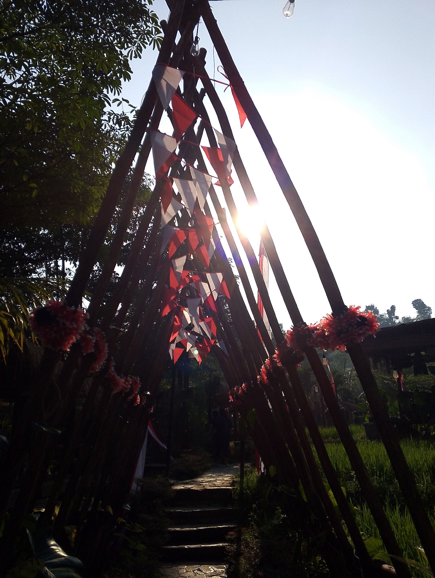 This image captures an outdoor scene with a triangular archway made of dark wooden poles, decorated with red and white pennant flags and red pom-pom garlands. Sunlight streams through the archway, creating lens flare and illuminating the sky. To the left of the archway, lush green trees are visible, their leaves dark against the bright sky. A stone pathway with steps leads through the archway and into a dimly lit area. To the right of the archway, a vibrant green rice field stretches out, bordered by trees in the distance. The overall impression is of a festive and natural setting, possibly a pathway leading to an event or a scenic viewpoint in Babakan Madang, Indonesia.