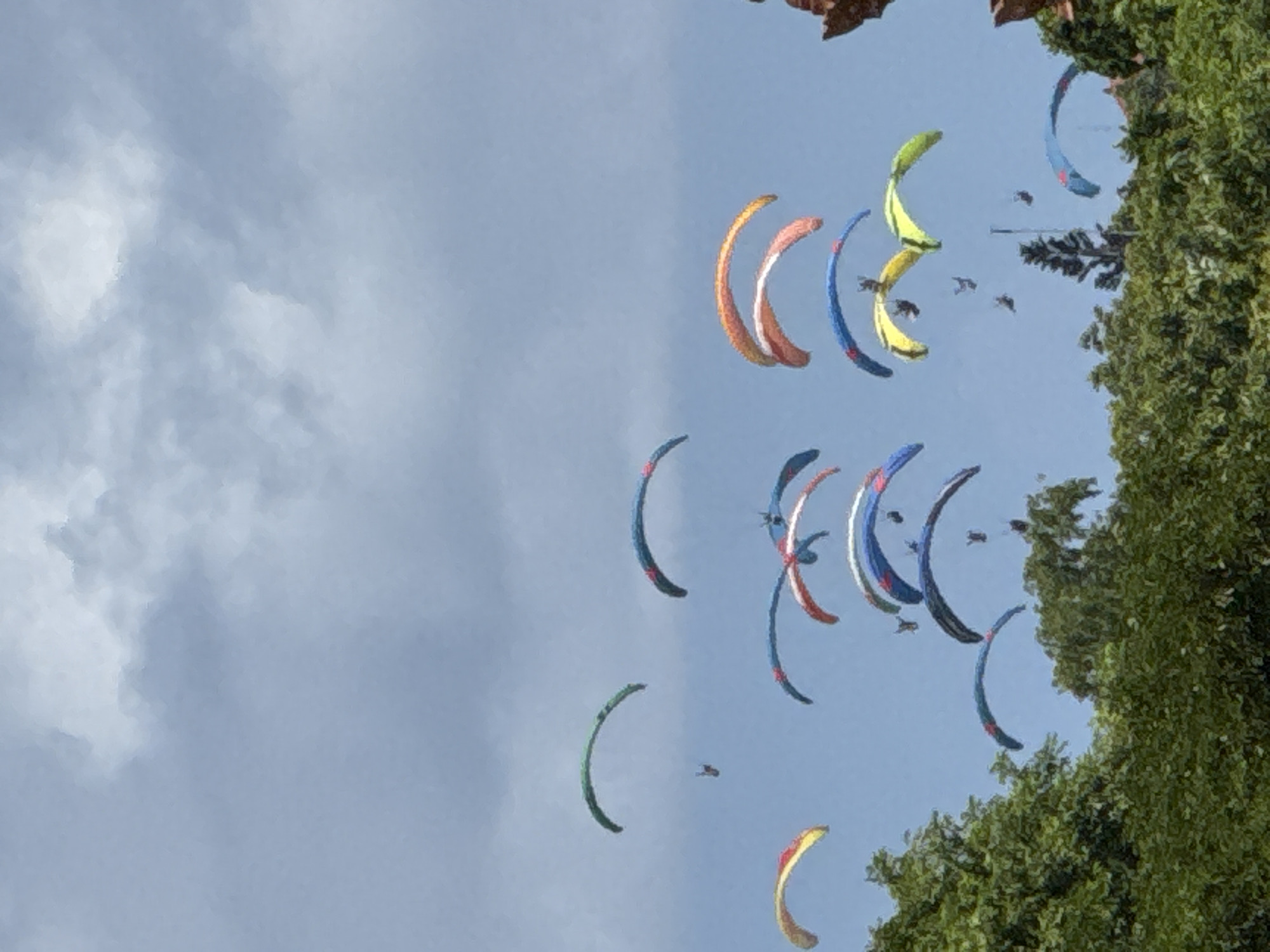 The image depicts numerous paragliders in the sky above Unknown, Indonesia. The sky is partially obscured by clouds. Trees are visible on the right side of the frame, and some buildings are barely visible. The paragliders are of various colors. Several small objects, possibly birds or insects, are visible in the sky near the paragliders.