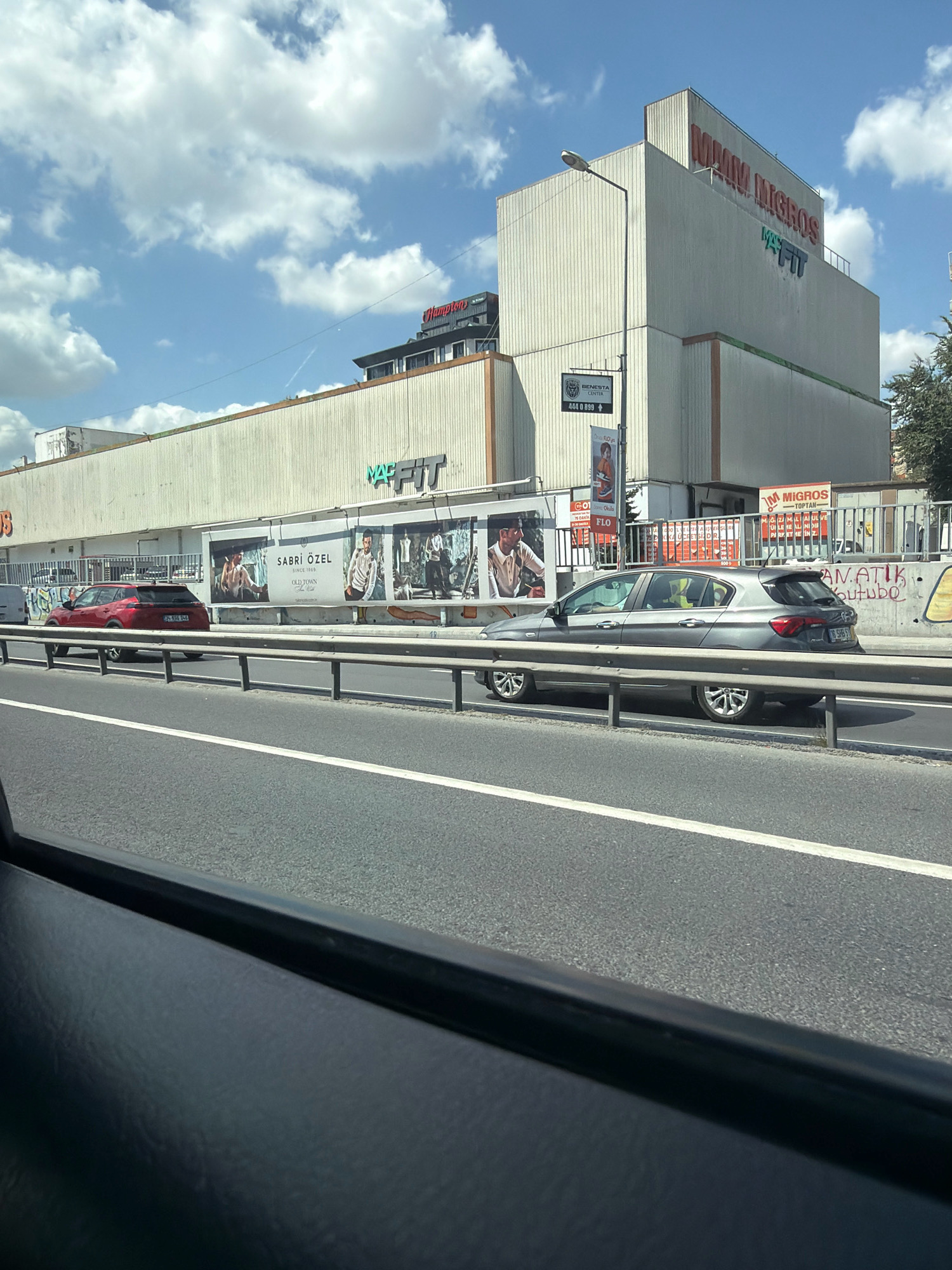 A high-angle, eye-level, medium shot captures a street scene in Güngören, Turkey, under a partly cloudy sky. The foreground shows a car's interior, with a dark dashboard and a portion of a car window on the left. Beyond the window, a multi-lane asphalt road runs horizontally across the frame. A metal guardrail separates the road from a sidewalk or elevated area.
On the far side of the road, a large, multi-story commercial building dominates the background. The building's facade is a pale, textured material, with some sections appearing to be corrugated. Large, illuminated signs are visible on the building. One prominent sign displays "MAM MIGROS" in red lettering, and below it, "MAC FIT" in green and blue.
Further along the building's facade, large advertisements are displayed. These appear to be billboards featuring models, clothing, and text that includes "SABRI OZEL SINCE 1969" and "OLD TOWN".
Several vehicles are visible on the road. A red SUV is parked to the left of the frame, with its license plate partially visible. A silver sedan is in motion, passing in front of the building and its advertisements. Another vehicle, a white van, is also visible on the road further in the distance.
The environment suggests an urban setting with commercial activity. The buildings, road, and advertisements are characteristic of a developed city area. The sky is a bright blue with scattered white clouds, indicating daylight. The overall impression is a snapshot of everyday life on a busy street in Güngören.