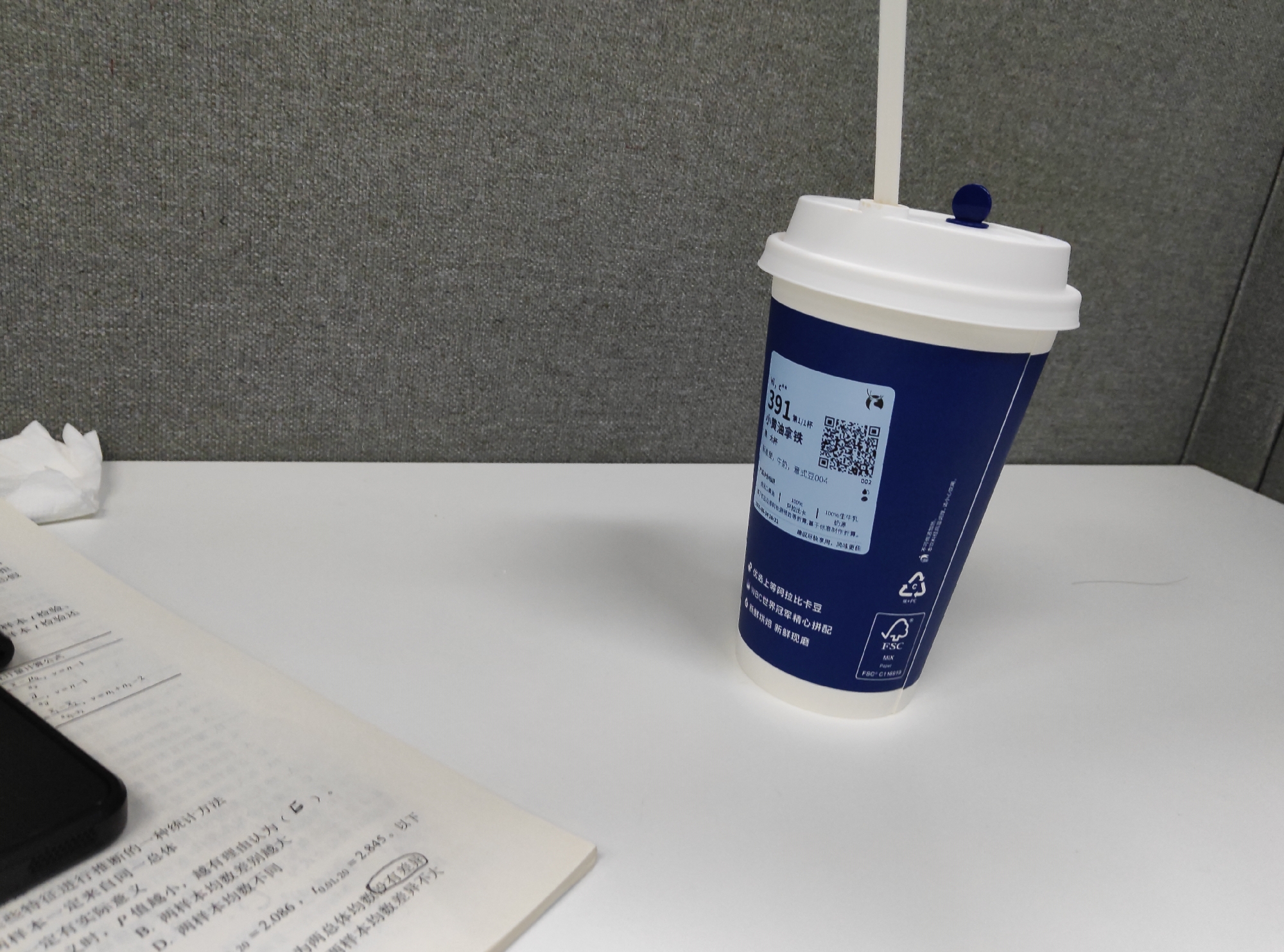 This media file captures a close-up, slightly tilted view of a blue and white disposable coffee cup with a straw, positioned on a white surface. The cup is adorned with text and a QR code, featuring the number "391" prominently and the words "小黄油拿铁" (Little Butter Latte) and "大杯" (Large Cup). Other visible text includes ingredient information, a QR code, and a FSC (Forest Stewardship Council) certification logo. To the left of the cup, a portion of an open book with text in Chinese is visible, along with a corner of a smartphone. In the background, a textured, grey fabric partition is present, suggesting an office or cubicle environment. The lighting appears to be artificial, indicative of an indoor setting, and there are no clear indicators of the time of day or weather conditions. The scene conveys a sense of a casual moment, perhaps a break at work, with the coffee cup and book suggesting activity related to study or work.