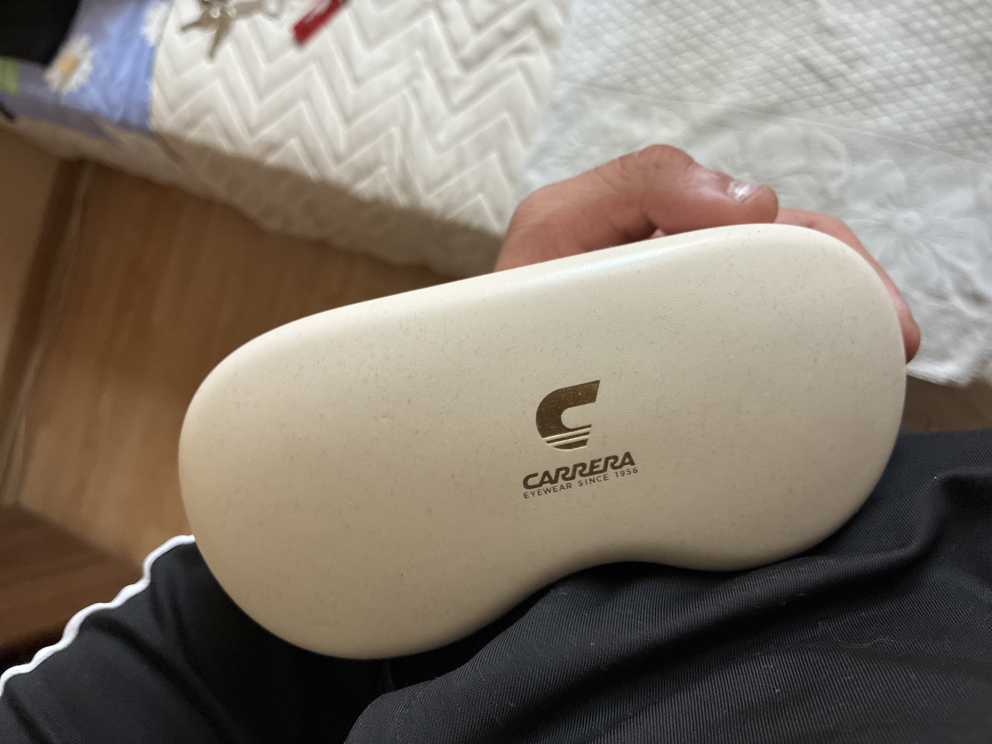A close-up, slightly overhead indoor shot shows a person holding an off-white or beige eyeglasses case on their lap. The case has a subtly textured surface and features the dark brown "CARRERA" logo, which includes a distinct stylized 'C' symbol and the text "EYEWEAR SINCE 1956." The person's hand, with a light skin tone, is visible gripping the case. The person is wearing black clothing, possibly athletic shorts or pants, distinguished by a white wavy stripe on the side.
In the blurred background, a domestic setting is evident. Parts of a bed or sofa are covered with patterned bedding; one section is white with a prominent zigzag pattern, while another segment is blue with a lighter floral design. A small set of keys, including one with a red accent, rests on this bedding. A light brown wooden surface, likely flooring or furniture, is also visible in the upper left. The lighting suggests it is daytime. No specific activities or emotions are discernible. The image itself does not provide explicit visual cues for the location Varna, Bulgaria.