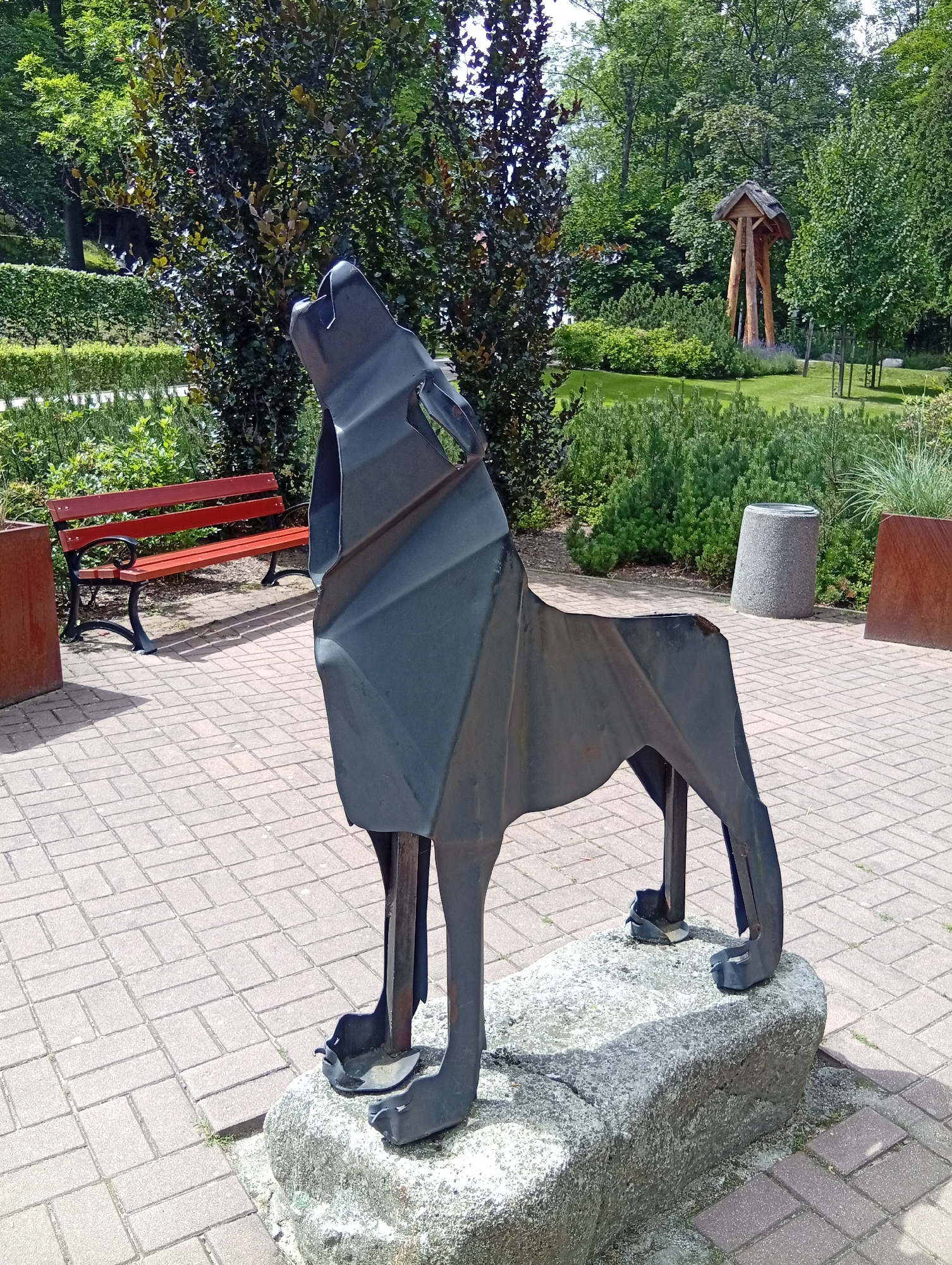 A dark metal sculpture of a wolf, crafted from angular pieces of metal, stands on a rough grey stone in a park. The wolf is depicted in a howling pose, with its head tilted upwards. The sculpture is positioned on a paved walkway made of light brown brick pavers arranged in a herringbone pattern. To the left of the sculpture, a red wooden bench with black metal legs is visible. In the background, there are various trees and shrubs, with a larger, more elaborate wooden structure with a peaked roof standing further back in a grassy area. The scene appears to be outdoors on a sunny day, with shadows cast by the sculpture and other elements. This sculpture is located in the Karpacz City Park in Karpacz, Poland.