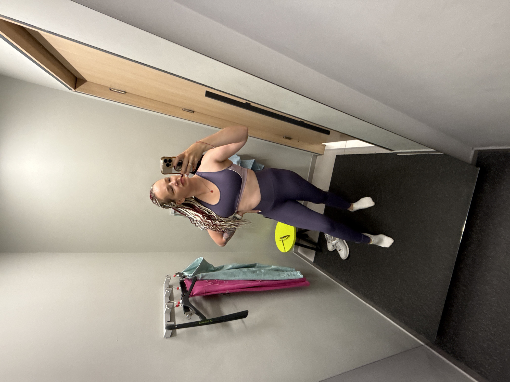 A woman is taking a mirror selfie in a fitting room. She is wearing a purple sports bra and matching leggings. She has braided hair with streaks of red and white. In the background, there is a rack with pink and blue garments, a yellow stool with sunglasses on it, and a pair of white sneakers. The fitting room has grey walls and a black floor mat. A wooden door with a black handle is visible on the right side of the image.
