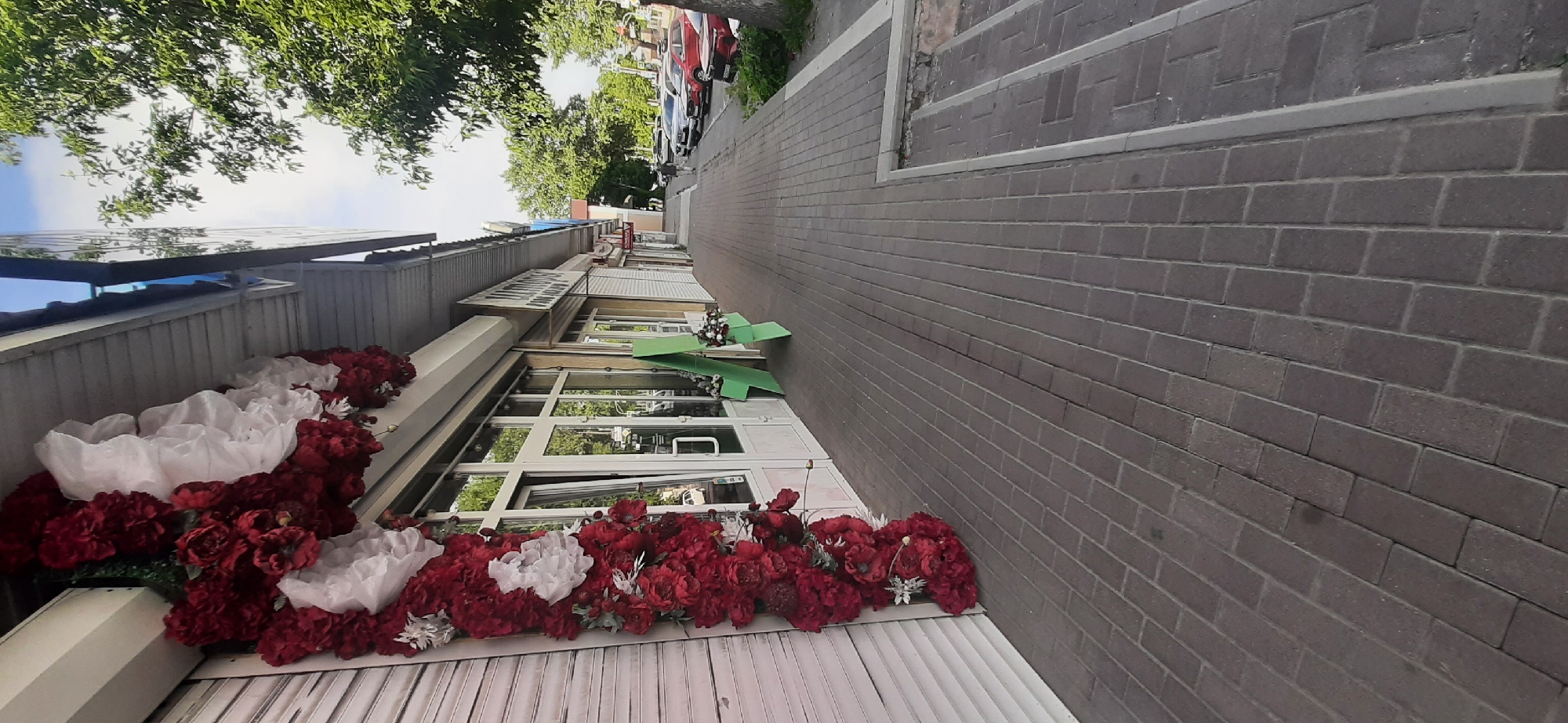 The image shows a row of shops with large windows. Artificial red and white flowers are displayed along the front of the shops. A brick sidewalk runs alongside the shops, and trees and cars are visible in the background. The scene appears to be outdoors during the daytime, with a bright sky visible through the trees. The overall setting suggests a commercial street or a pedestrian walkway next to a business district. There is no discernible activity or event taking place, and no visible text. The image is taken from a low angle, looking up towards the buildings and trees.