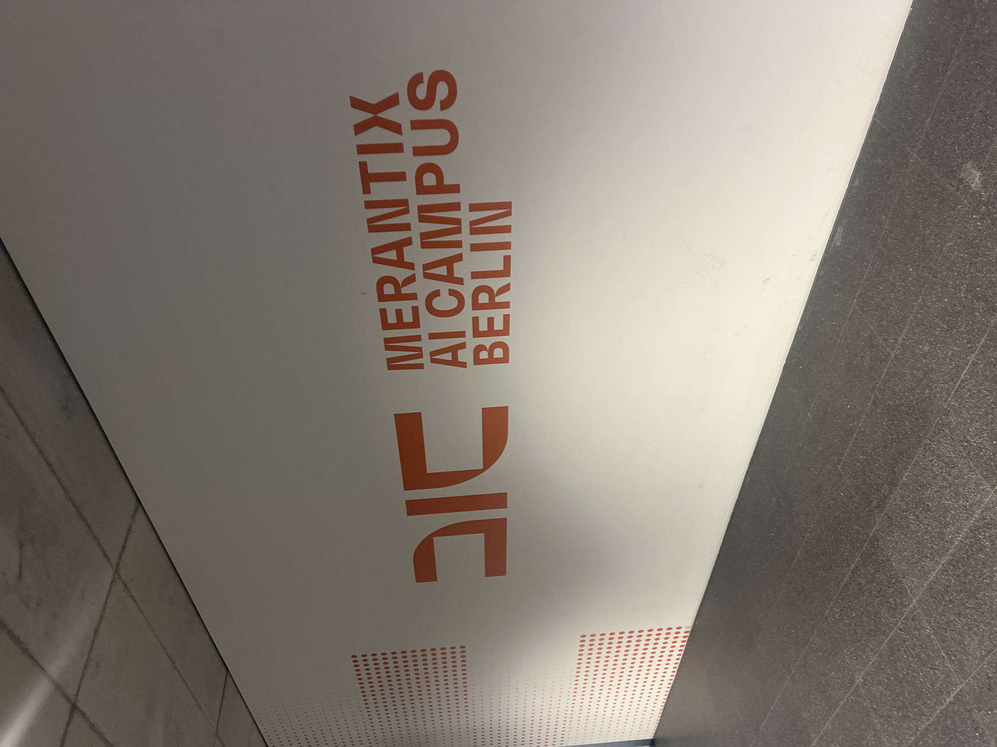 An orange logo and text are displayed on a white wall in a modern, indoor setting in Berlin, Germany. The logo, appearing to be abstract geometric shapes, is located to the left of the text. The text reads "MERANTIX AI CAMPUS BERLIN" in stacked lines. The wall has a textured pattern of red dots fading from dense to sparse on the left side, extending from the top to about a third of the way down the wall. The floor is dark, speckled tile, and the ceiling appears to be a concrete structure with visible lines. The lighting suggests an artificial light source, casting slight shadows. There are no people or animals visible in the image.