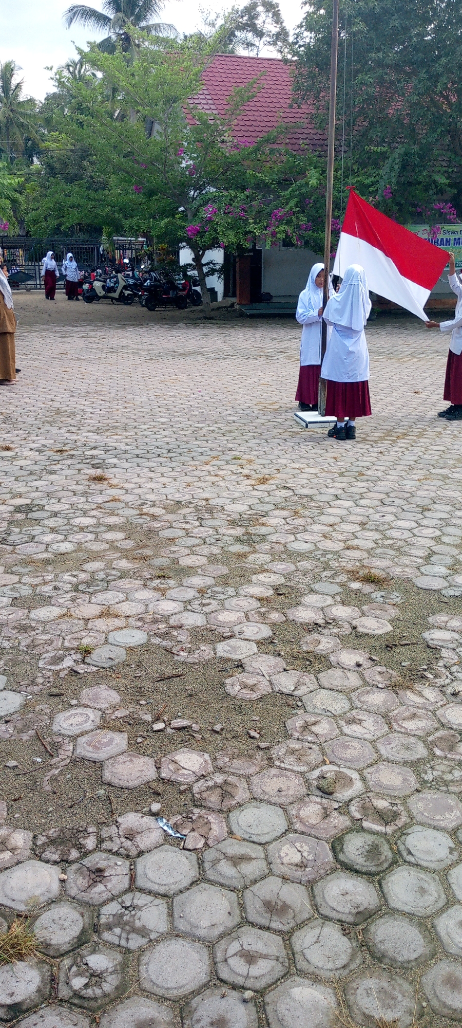 In Baratpayaitek, Indonesia, a group of female students in uniform stand near a flagpole in a paved outdoor area. The students, wearing white headscarves and maroon skirts, are participating in what appears to be a flag-raising ceremony. One student is holding the Indonesian flag, which is red and white, while others stand beside her. In the background, a building with a red roof is visible, surrounded by trees and vegetation. Several motorcycles are parked to the left of the frame, suggesting this location might be a school or community gathering space. The ground is paved with hexagonal tiles, some of which are cracked and covered with sand and debris.