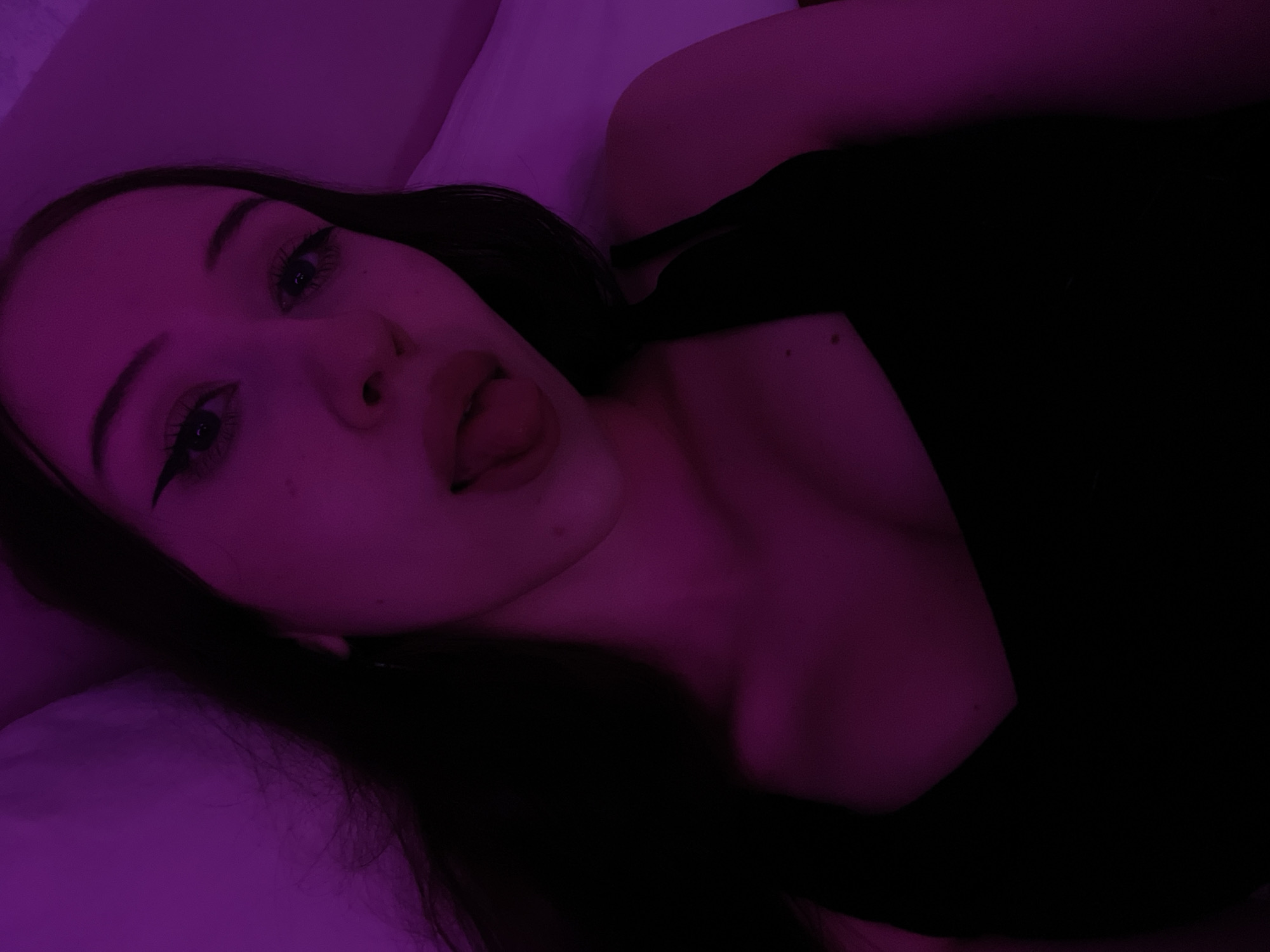 A young woman is captured in a close-up selfie against a purple-lit background. She is wearing a black tank top and has dark hair. Her tongue is sticking out, and she has winged eyeliner. The setting appears to be indoors, possibly a bedroom, with a pillow visible in the background. There is no indication of her location within Votkinsk, Russia.