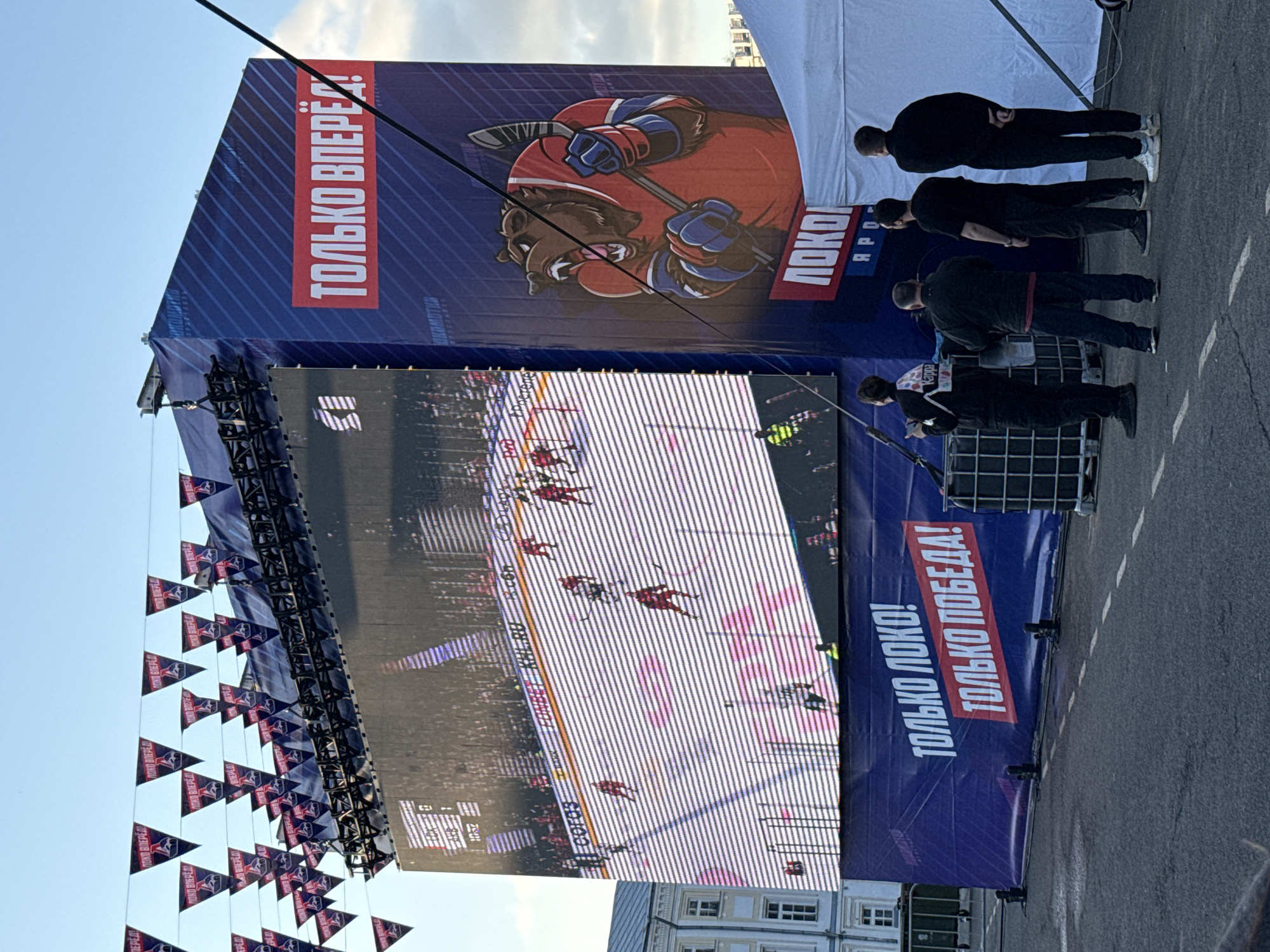The image shows a large outdoor screen displaying a hockey game. The screen is set up in a public area, possibly a street or square, with paving stones visible in the foreground. A banner above the screen features a cartoon image of a bear with a hockey stick, as well as the text "ТОЛЬКО ВПЕРЁД!" (Only Forward!) and "ЛОКО ЯРОГ." (Loco Yarog). There are also flags hanging from the structure supporting the screen. Three people are standing to the right of the screen, all wearing dark clothing. The environment suggests this is an event or public viewing of a hockey game. Based on the text on the banners and logos, it is located in Russia.