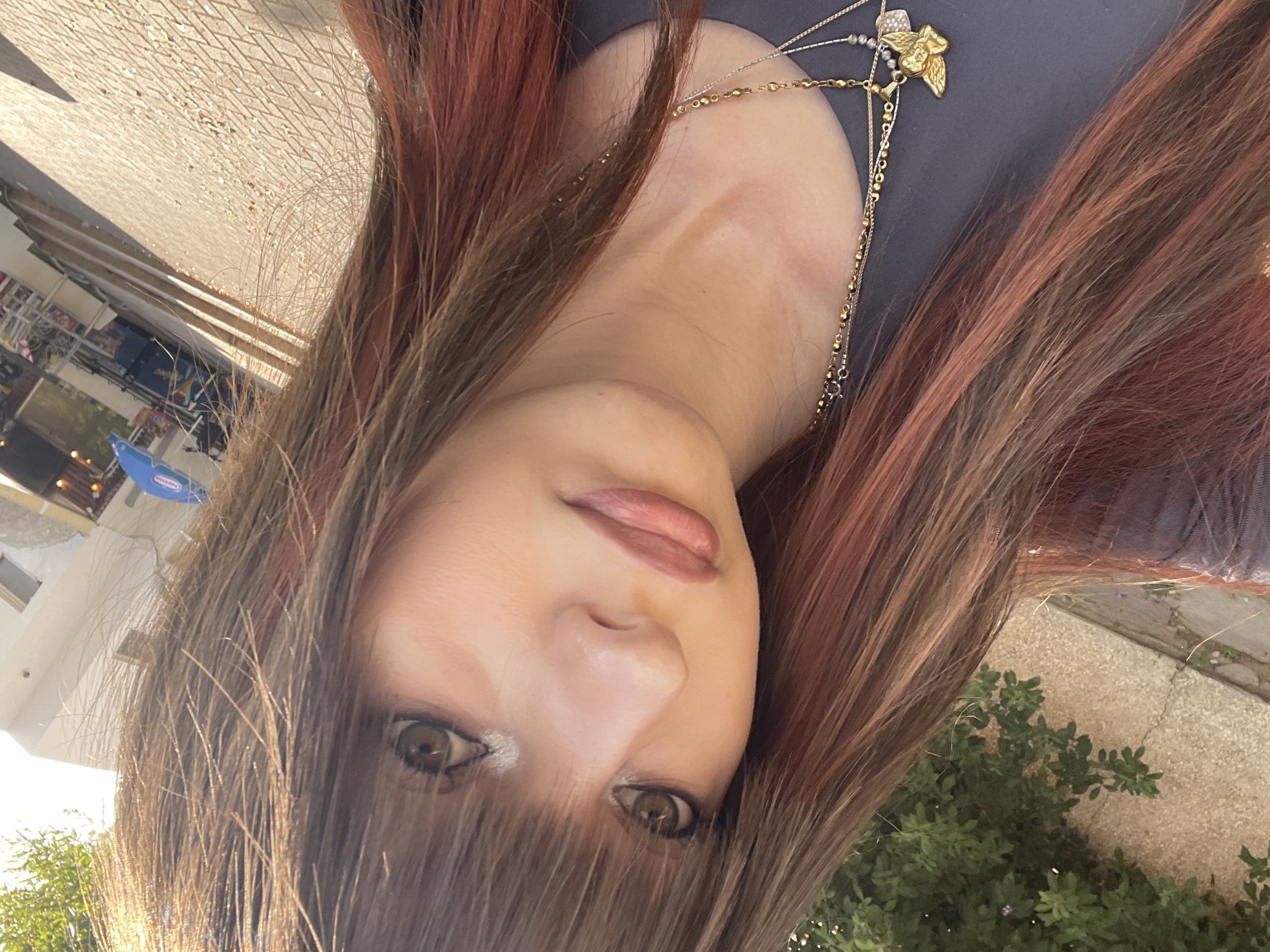 The image shows a close-up, angled selfie of a young woman with long, layered brown hair featuring subtle reddish-pink highlights. She has light-colored eyes with prominent eyelashes, light pink lipstick, and appears to be wearing a dark grey top layered with multiple delicate gold necklaces. One necklace has a small angel pendant. The background is slightly blurred, showing an outdoor urban environment with a paved walkway, greenery, and buildings in the distance, suggesting a sunny day. This scene is consistent with a location in Raanana, Israel, known for its urban parks and tree-lined streets.
