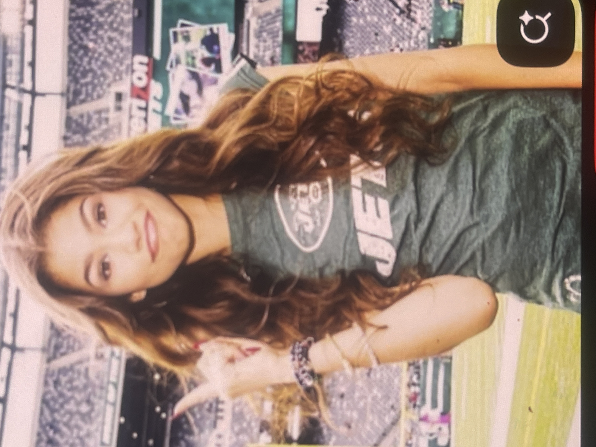 The image displays a young woman with long, wavy brown hair, smiling and looking directly at the camera. She is wearing a dark green t-shirt featuring the circular logo of the New York Jets and the word "JETS" prominently printed on the front. Her right hand is raised, with the index finger pointing forward, and she has multiple bracelets on her wrist.
The background depicts what appears to be a large sports stadium or arena. Visible elements include rows of seating for spectators, a vibrant green field, and advertising banners. The word "Verizon" is clearly legible on one of these banners. The lighting suggests it is daytime. The overall setting implies an outdoor event, possibly a football game or a fan gathering. The woman's expression conveys happiness and engagement. The image itself is rotated 90 degrees counter-clockwise.