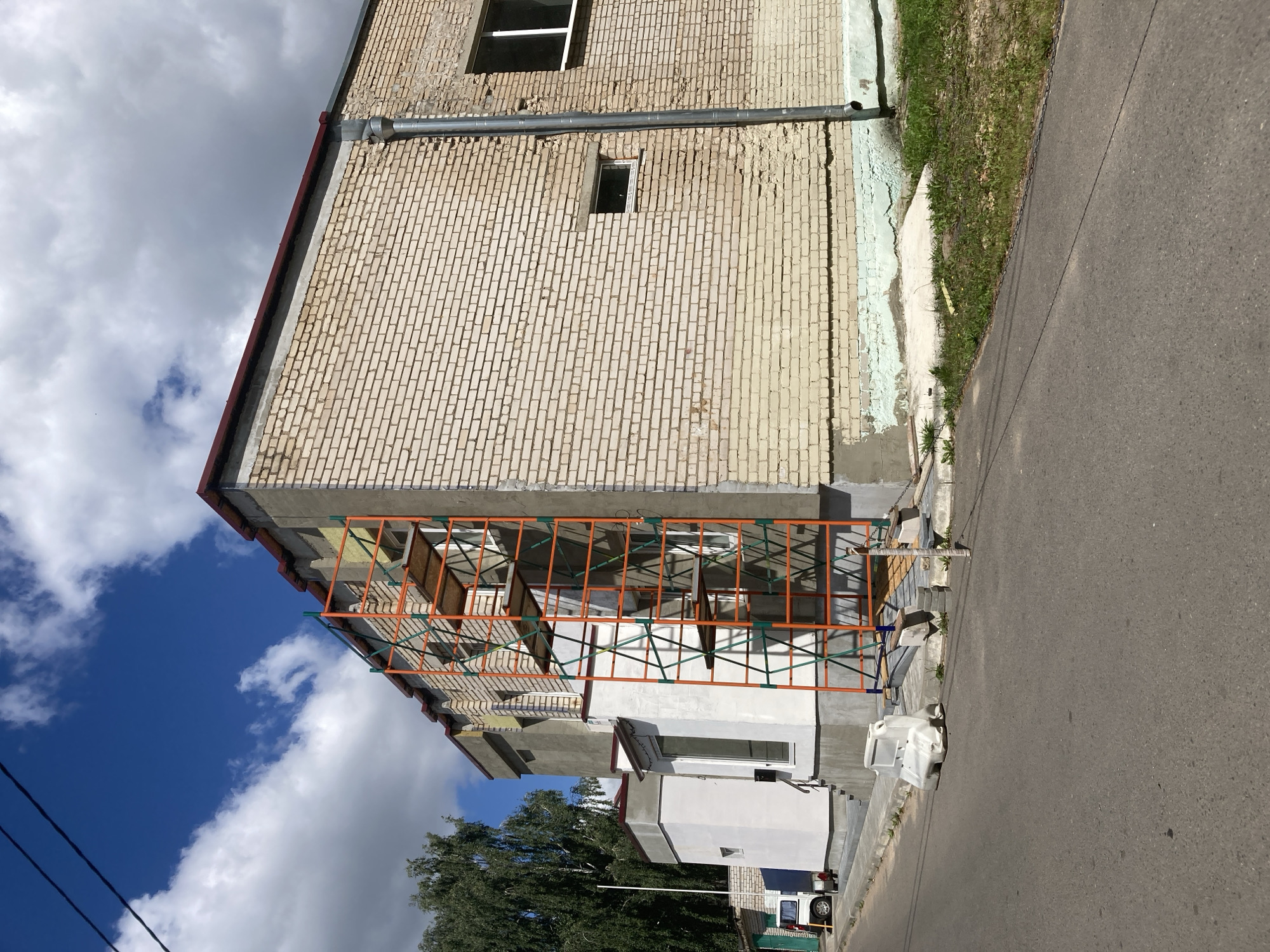 The image shows a light beige brick building undergoing renovation, featuring scaffolding on its side. The building has a dark red roofline and several windows, some of which are visible. A downspout runs vertically along the right side of the building. The area in front of the building appears to be a road or street, with a sidewalk and some grass. There is a white plastic water barrier on the sidewalk. In the background, a tall green tree can be seen. The sky above is blue with scattered white clouds. The scene is located in the city of Zhodzina, Belarus.