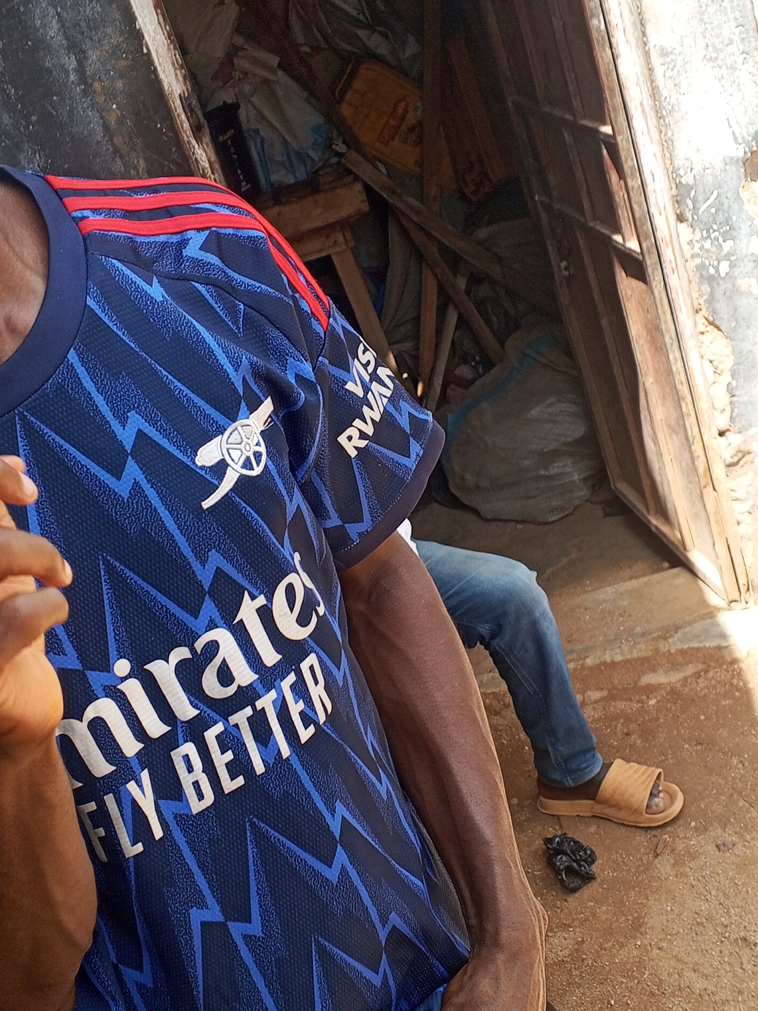 A person wearing a blue Arsenal soccer jersey with the Emirates logo is partially visible in the foreground. Red stripes are seen on the shoulder of the jersey. A person is seated behind them, wearing blue jeans and brown sandals. A small black plastic bag sits on the ground near their feet. They are positioned in the doorway of a building with weathered wooden beams and door frame. The inside of the building appears cluttered with various items, including bags and a wooden stool. The scene is likely in an outdoor environment, but it is difficult to determine if the scene is really located in Gwong, Nigeria.