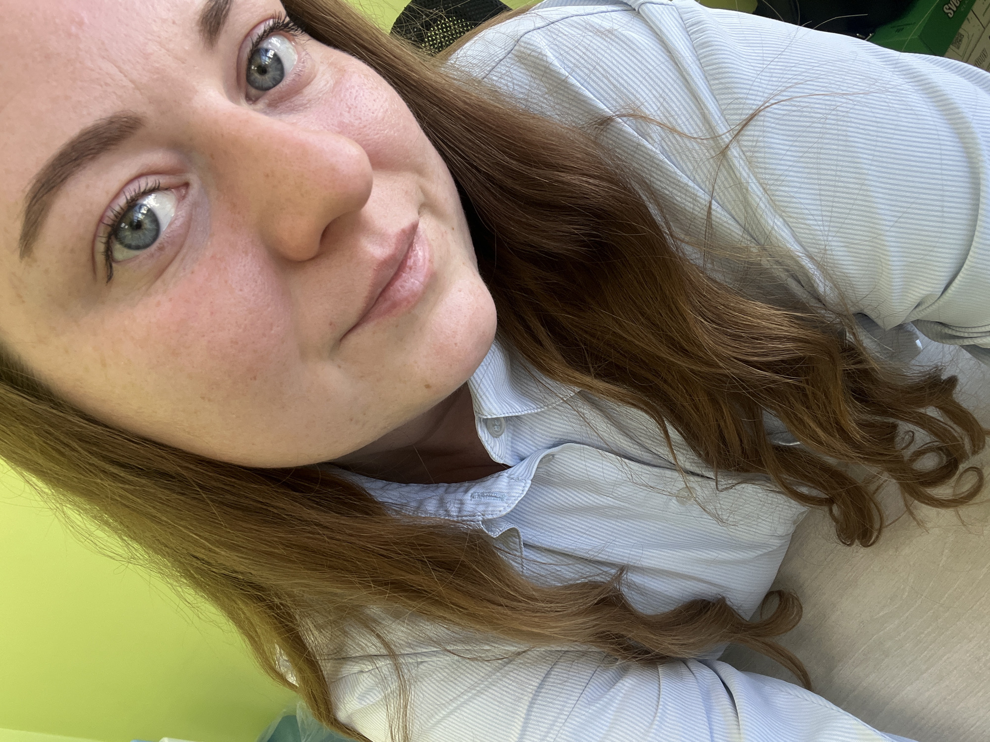 A close-up, angled selfie depicts a fair-skinned woman with reddish-brown hair and blue eyes. She is wearing a light blue striped shirt. Her facial features are clear, and she has visible freckles. The background consists of a light green wall and a glimpse of what appears to be a desk surface on the lower right. The exact location within Klin, Russia, cannot be determined from this image.