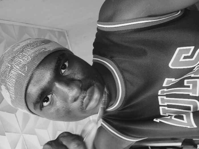 A black and white, head-and-shoulders portrait of a young male subject is captured in an indoor setting within Bukuru, Nigeria. The subject, featuring dark skin and a faint mustache, looks directly into the camera. He wears a patterned knit beanie and a dark basketball jersey with "BULLS" partially visible in white lettering with red outlines on the chest. The jersey's neckline and armholes have striped trim in white and red. The subject's left hand is partially visible on the left side of the frame, indicating a self-portrait. The background consists of a geometric textured panel with a repeating triangular or pyramidal pattern on the upper left and right, contrasted by a plain, lighter-toned wall section.