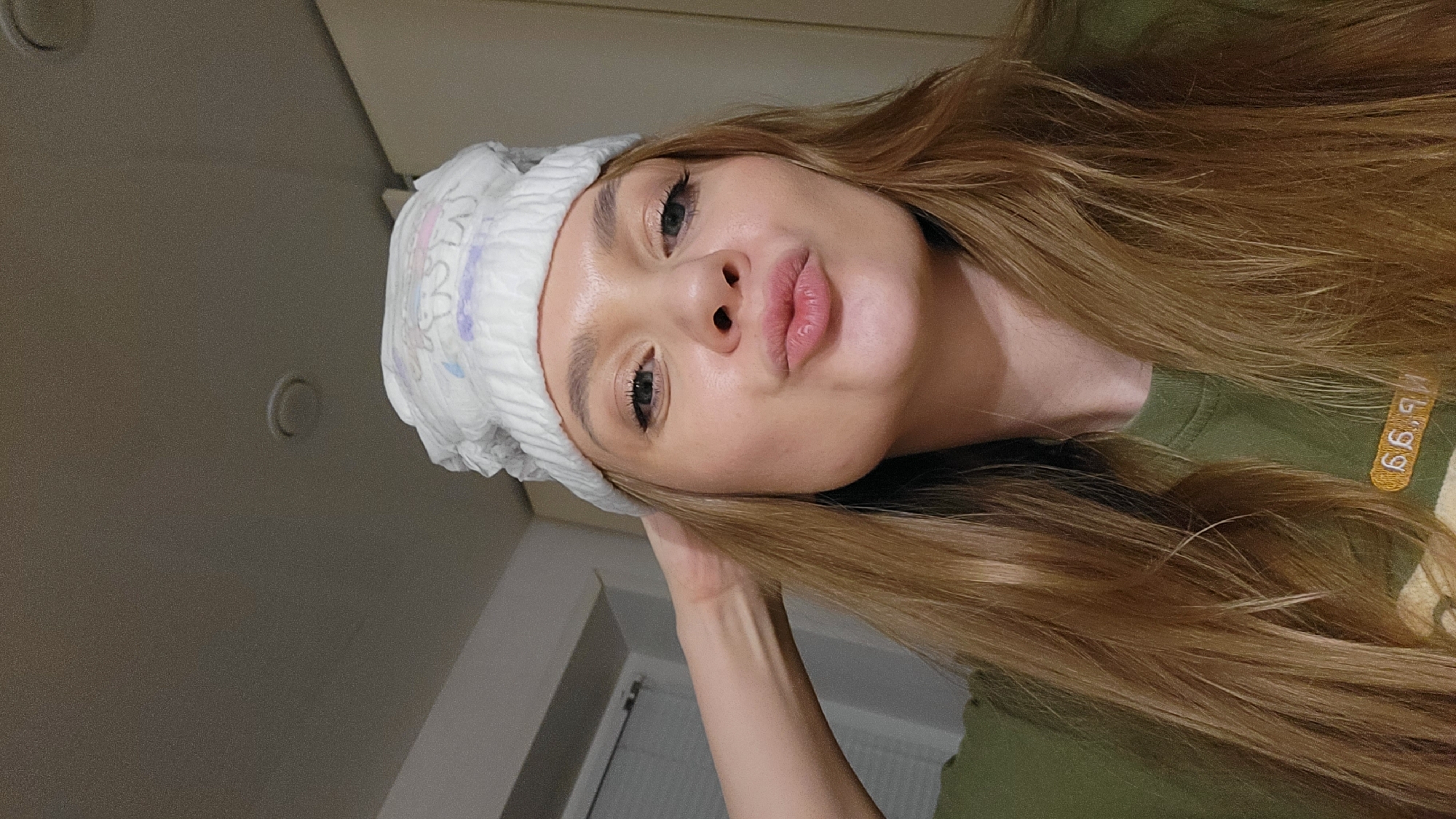 A woman with long, light brown hair is posing for a selfie. She is wearing an olive green t-shirt and what appears to be a diaper on her head. The background is a lightly colored wall with a small, round object on it. A doorway is visible in the background. There is nothing in the image to suggest a location in the city of Chaikovsky, Russia.