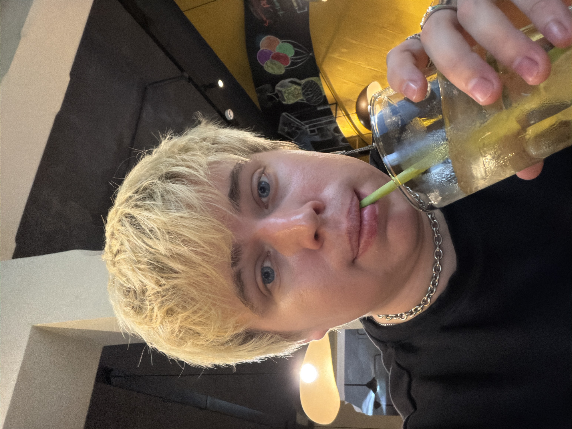 A person with short, light blonde hair and blue eyes, wearing a black short-sleeved top and a silver-colored chunky chain necklace, is seen in a medium close-up, drinking from a clear glass with a green straw. The glass contains a light amber liquid with ice cubes and a green stalk (likely lemongrass). Rings are visible on the thumb and index finger of the person's right hand, which holds the glass. The setting is indoors, appearing to be a cafe or restaurant located in Thủ Đức, Vietnam. The background on the right features a vibrant yellow wall beneath a dark, possibly chalkboard, section adorned with white chalk drawings of a stylized woman in a traditional dress and a cluster of colorful balloons, along with some indistinct white text. On the left, a large, elongated yellow pendant light fixture with a visible bulb glows brightly behind the person's head, and other dark pendant lights and a dark ceiling are visible further back.