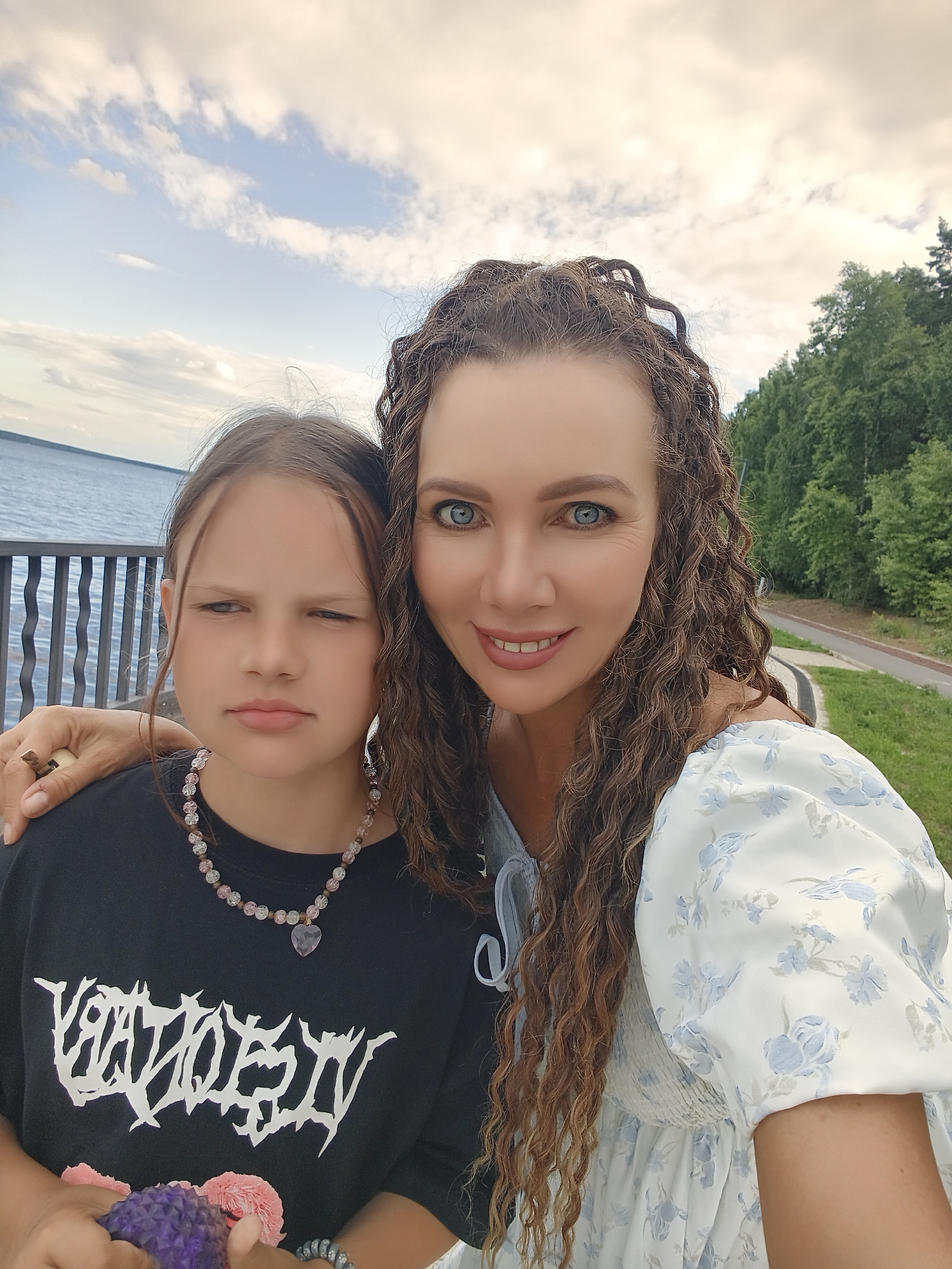 This media file is a selfie taken outdoors with two people in the foreground, a woman and a young girl. The woman is on the right, smiling widely, and has long, curly brown hair and blue eyes. She is wearing a white dress with a blue floral pattern. The young girl is on the left, looking away from the camera with a neutral expression. She has brown hair and is wearing a black t-shirt with a white graphic design. She is also wearing a beaded necklace with a heart-shaped pendant. The woman has her arm around the girl.
In the background, there is a body of water on the left, likely a river or lake, with a dark railing in front of it. On the right, there are trees and a paved path, suggesting a park or waterfront area. The sky is partly cloudy, indicating daytime. The overall mood of the image is casual and familial. The visible text on the girl's shirt appears to be in a stylized font, but it is not clearly legible. The setting appears to be a public outdoor space, possibly a promenade or park, consistent with the provided location context of Chaikovsky, Russia.