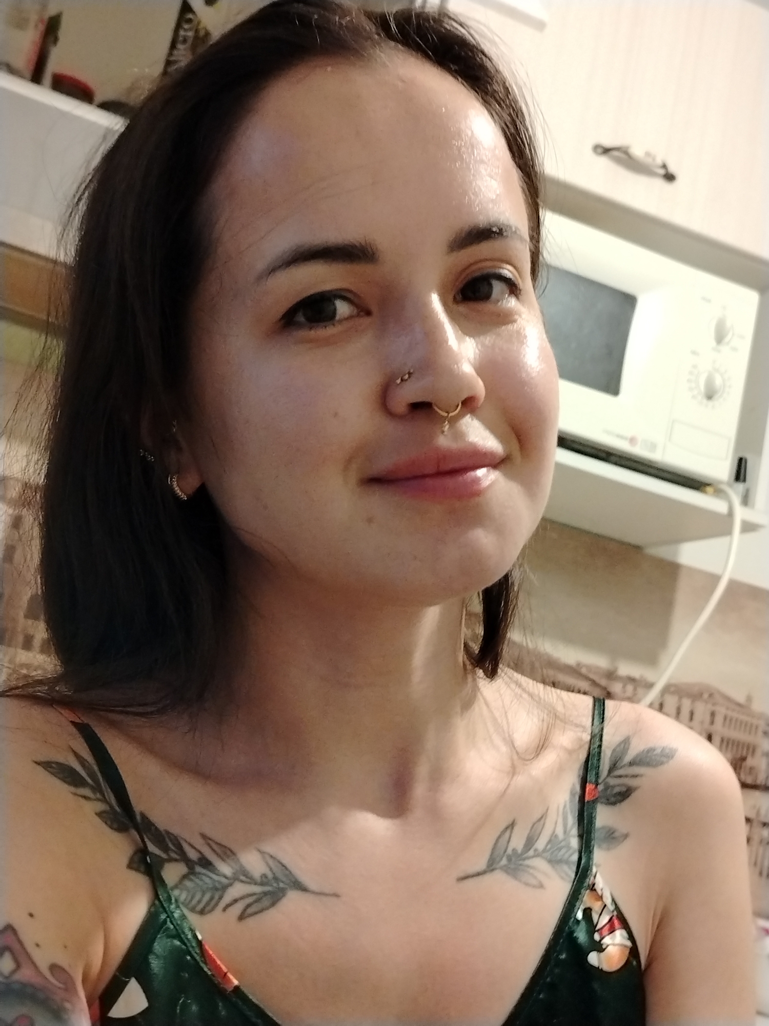 A young woman is centered in the frame, posing for a selfie in what appears to be a kitchen. She has dark hair, light skin, and multiple piercings in her nose. Her attire includes a dark-colored camisole with thin straps and a floral pattern. Two leafy branch tattoos are visible on her upper chest and shoulders.
Behind her, a white microwave sits on a shelf, and a wallpaper depicting a cityscape is partially visible. The wallpaper hints at architecture possibly from the city of Noviy. There is not enough information to verify that this location is in Russia. The lighting suggests an indoor environment, possibly artificially lit.