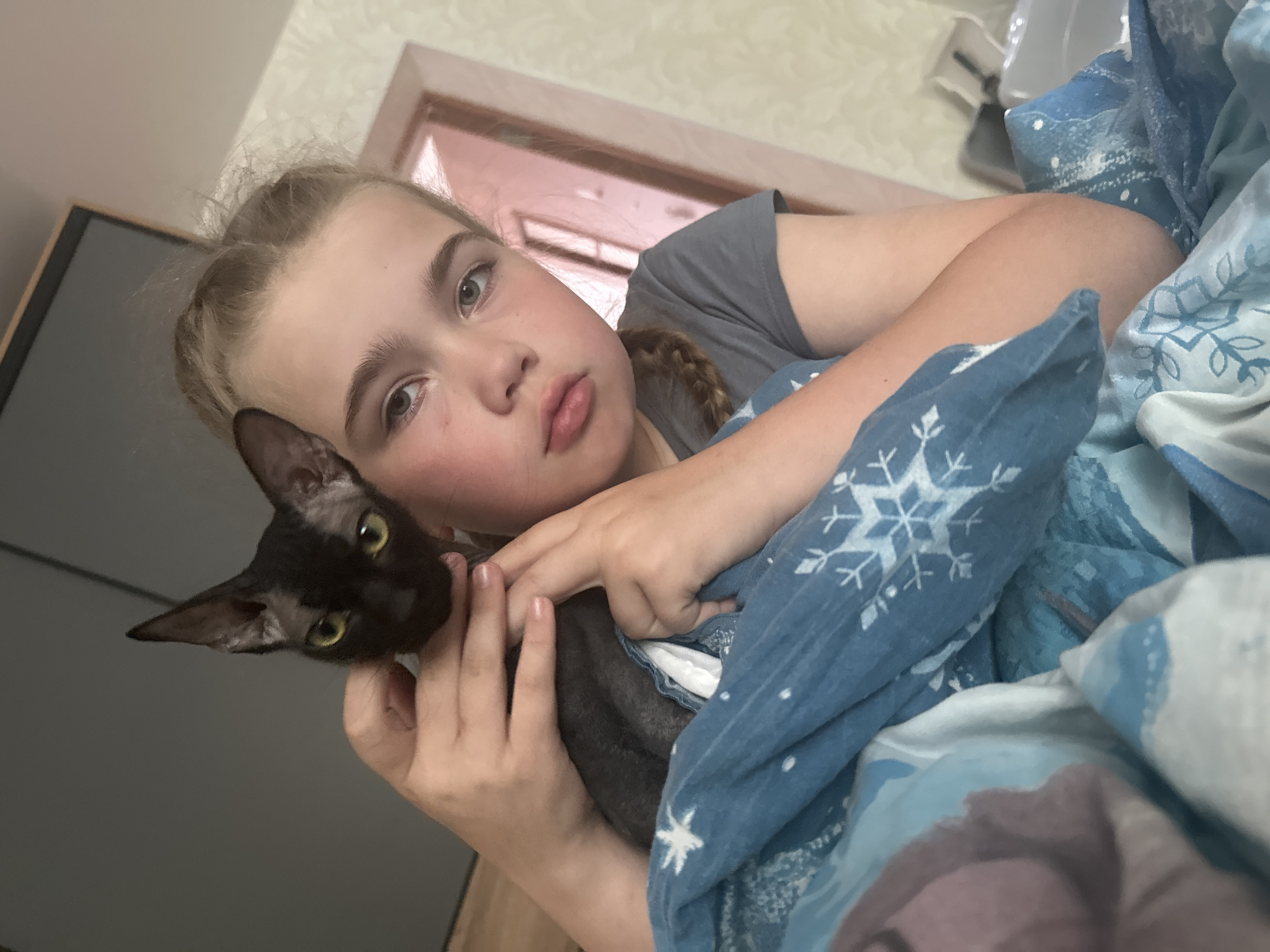 In a domestic indoor setting, a young Caucasian girl with blonde hair pulled back in a braid and grey eyes is holding a black Sphinx cat. The girl is wearing a grey t-shirt and is partially covered by a blue blanket with a snowflake pattern. The cat is black with large ears and yellow eyes, and it is resting on the girl's chest and shoulder. The background consists of a wall with textured wallpaper and a doorway. The scene is likely within a residential apartment in Chaikovsky, Russia, given the context provided.