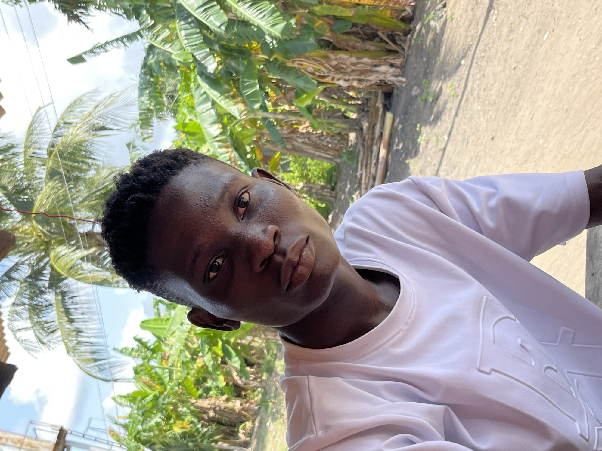 A medium close-up, waist shot of a Black African male in Badagry, Nigeria, looking at the camera. The man has short, dark hair and is wearing a white t-shirt with what appears to be embossed lettering on the front. The environment shows a mix of tropical vegetation, including banana trees, suggesting a rural or semi-rural area. The sky is partially visible, suggesting a bright, possibly sunny day. The location appears to be either on or near a dirt road or path.