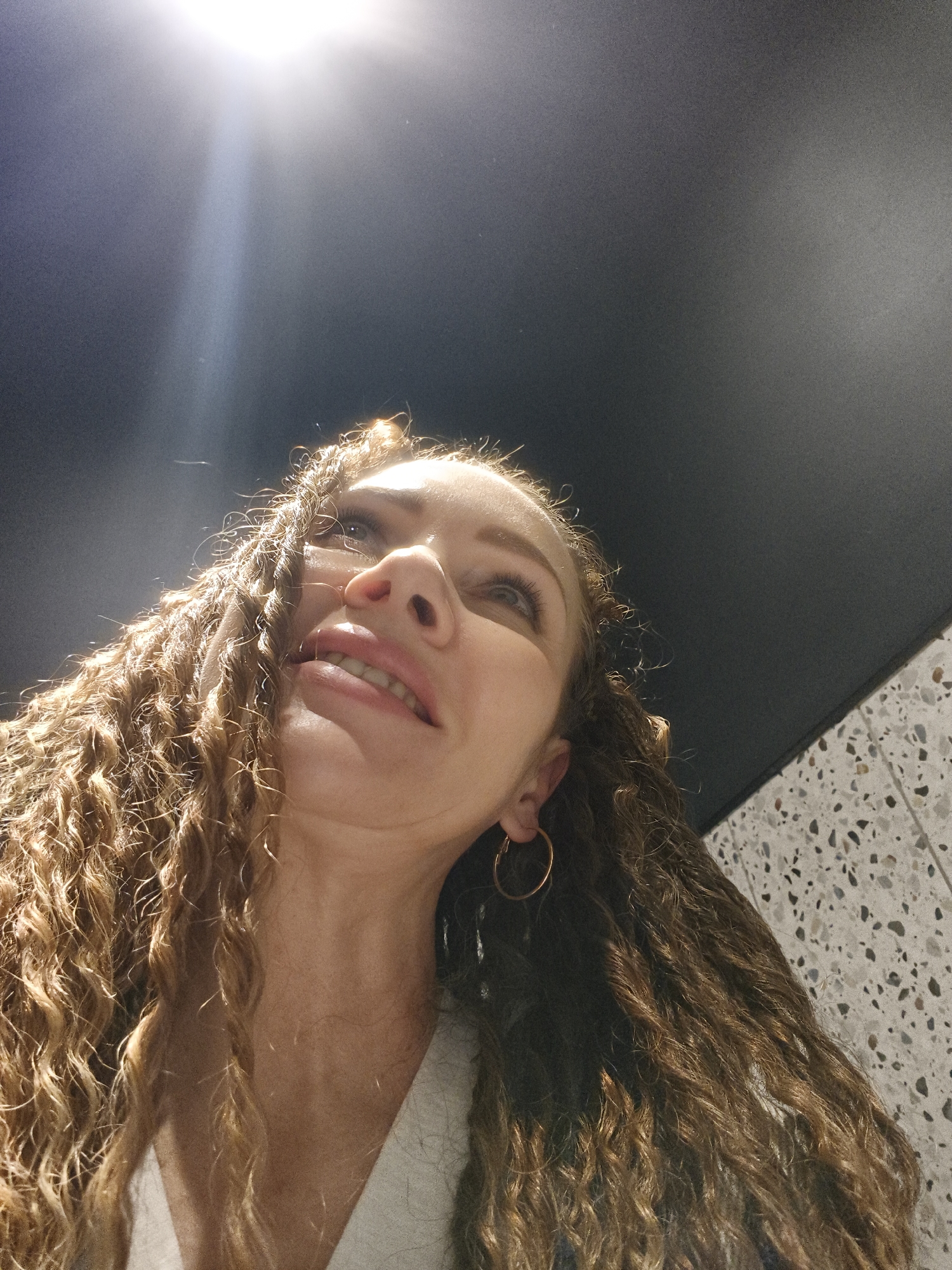 A low-angle view shows a woman with long, wavy, light brown hair looking upwards. She is wearing a gold hoop earring on one ear. The background is a dark, seemingly smooth surface with a bright light source at the top, along with a section of a patterned surface, possibly a wall. The location is Unknown, Unknown.
