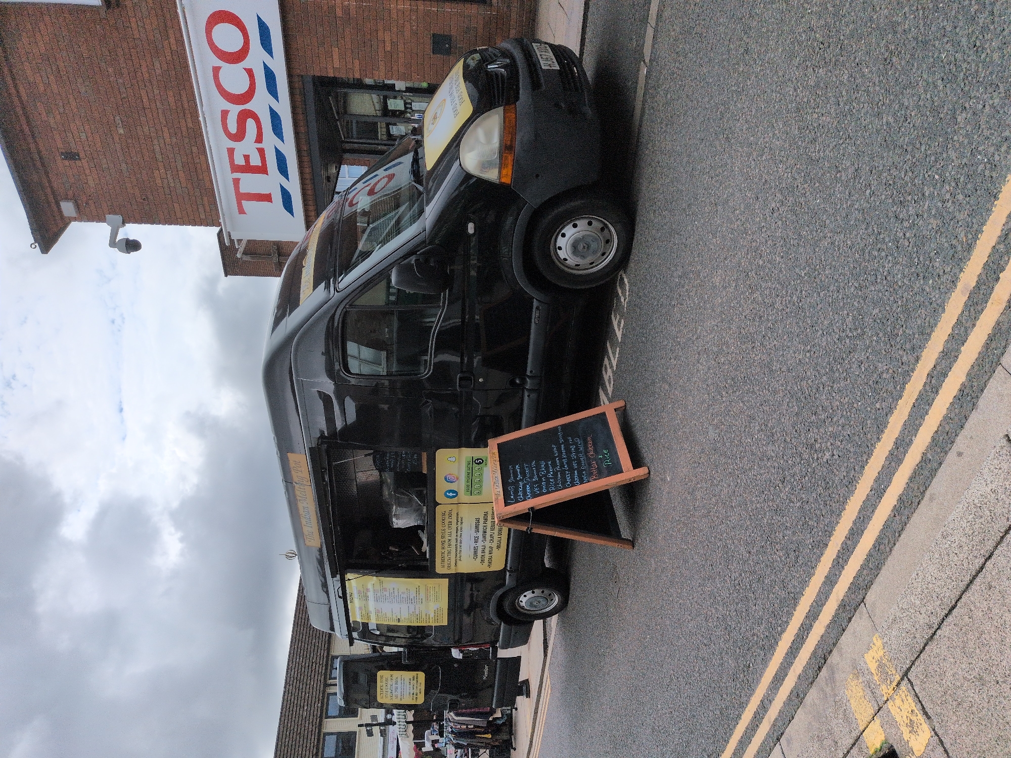 The image depicts a black food truck named "The Indian Melting Pot" parked on a street in Telford, United Kingdom. The truck is positioned in front of a Tesco supermarket, indicated by a prominent "TESCO" sign on the brick building. A black A-frame sign is placed in front of the food truck, displaying a menu. The street has double yellow lines painted on it, suggesting it's a restricted parking area. The scene appears to be outdoors, under a cloudy sky. There are no people clearly visible interacting with the food truck or the environment. Clothing racks and other items are visible to the left of the food truck, suggesting a market or outdoor retail setup. A security camera is mounted on the Tesco building. The overall impression is of a typical street scene with a food vendor operating near a supermarket.