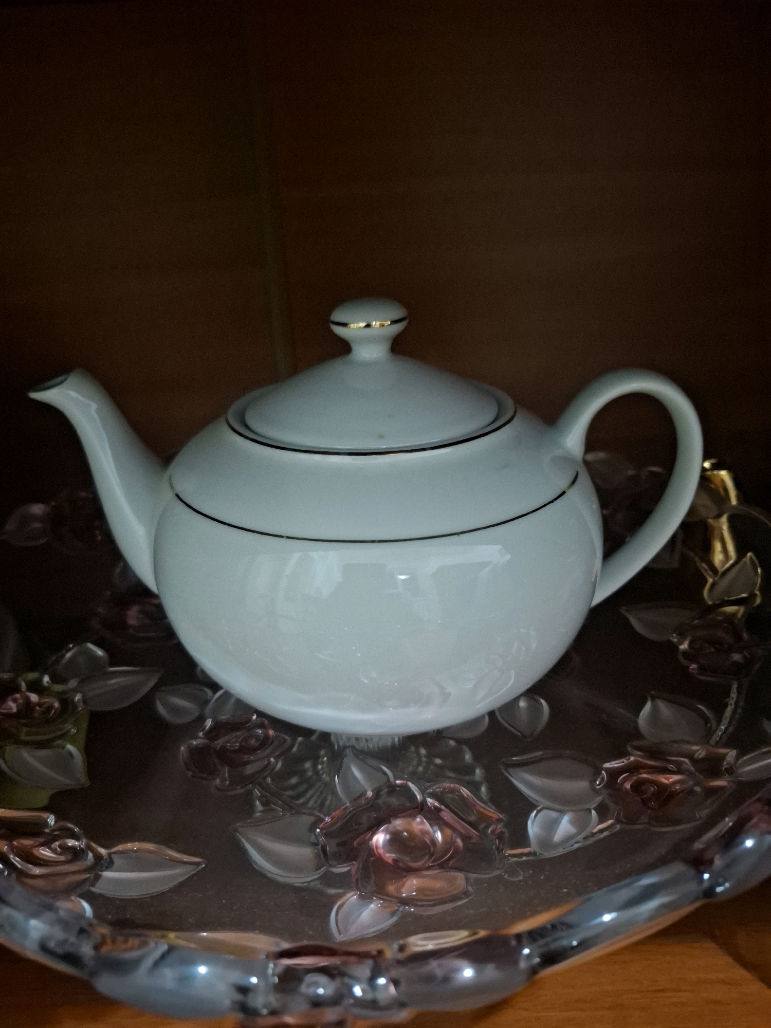 A white ceramic teapot with gold trim sits on a decorative glass plate featuring rose and leaf motifs. The teapot has a spout, a handle, and a lid with a small gold knob. The plate is translucent with red and clear glass embellishments. The background is a dark brown, wooden surface, suggesting the items are on a shelf or table. The lighting is dim, creating a moody atmosphere. This image was taken indoors, likely in a home or a display cabinet, and there is no indication of its location in Almaty, Kazakhstan. There are no people or actions depicted in the image.
