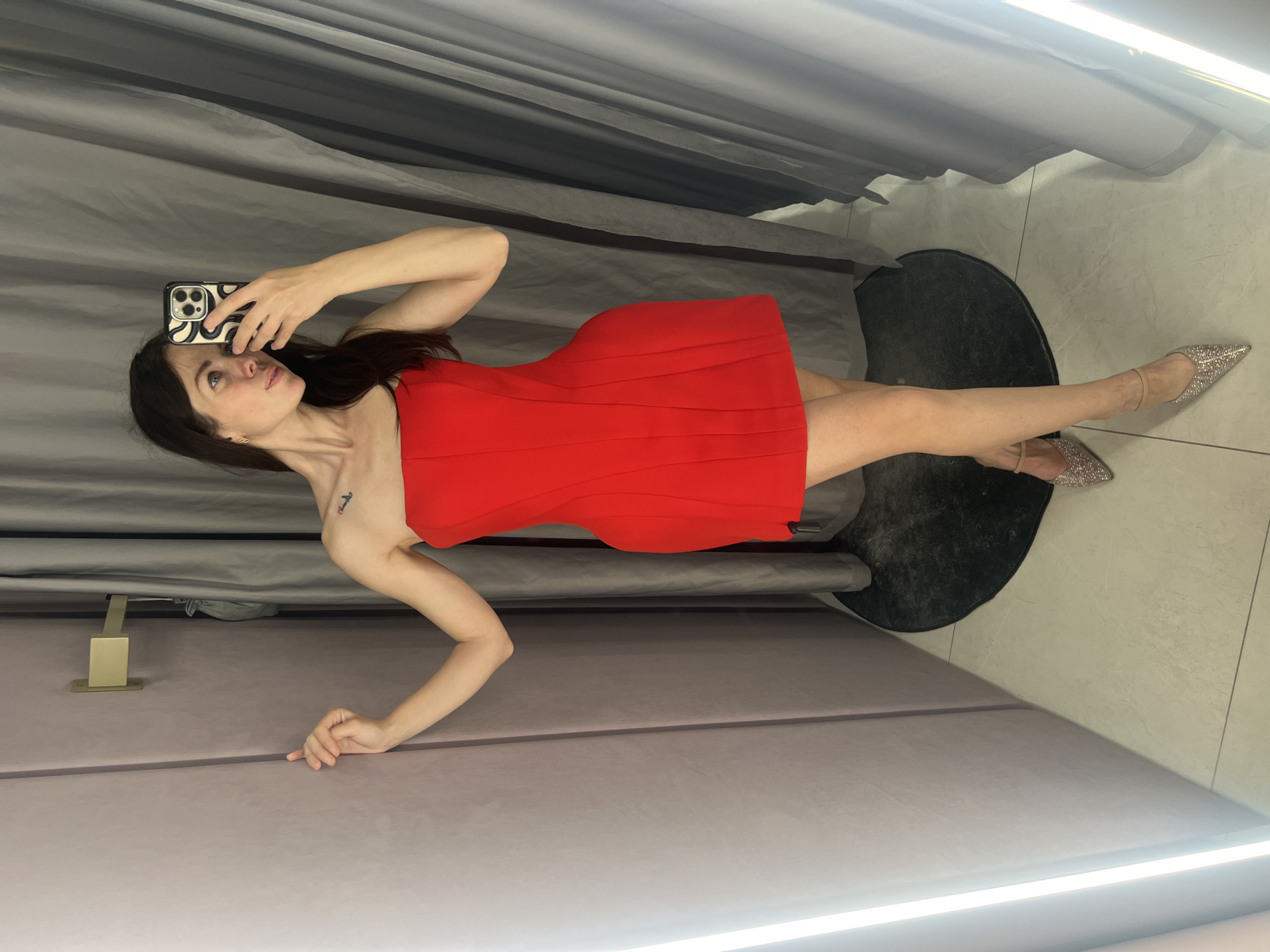 In a fitting room, a woman with dark hair stands in front of a mirror, taking a selfie. She is wearing a strapless red dress and sparkly pointed-toe heels. The fitting room has light gray curtains and a pale pink wall. A gold hook is mounted on the wall.