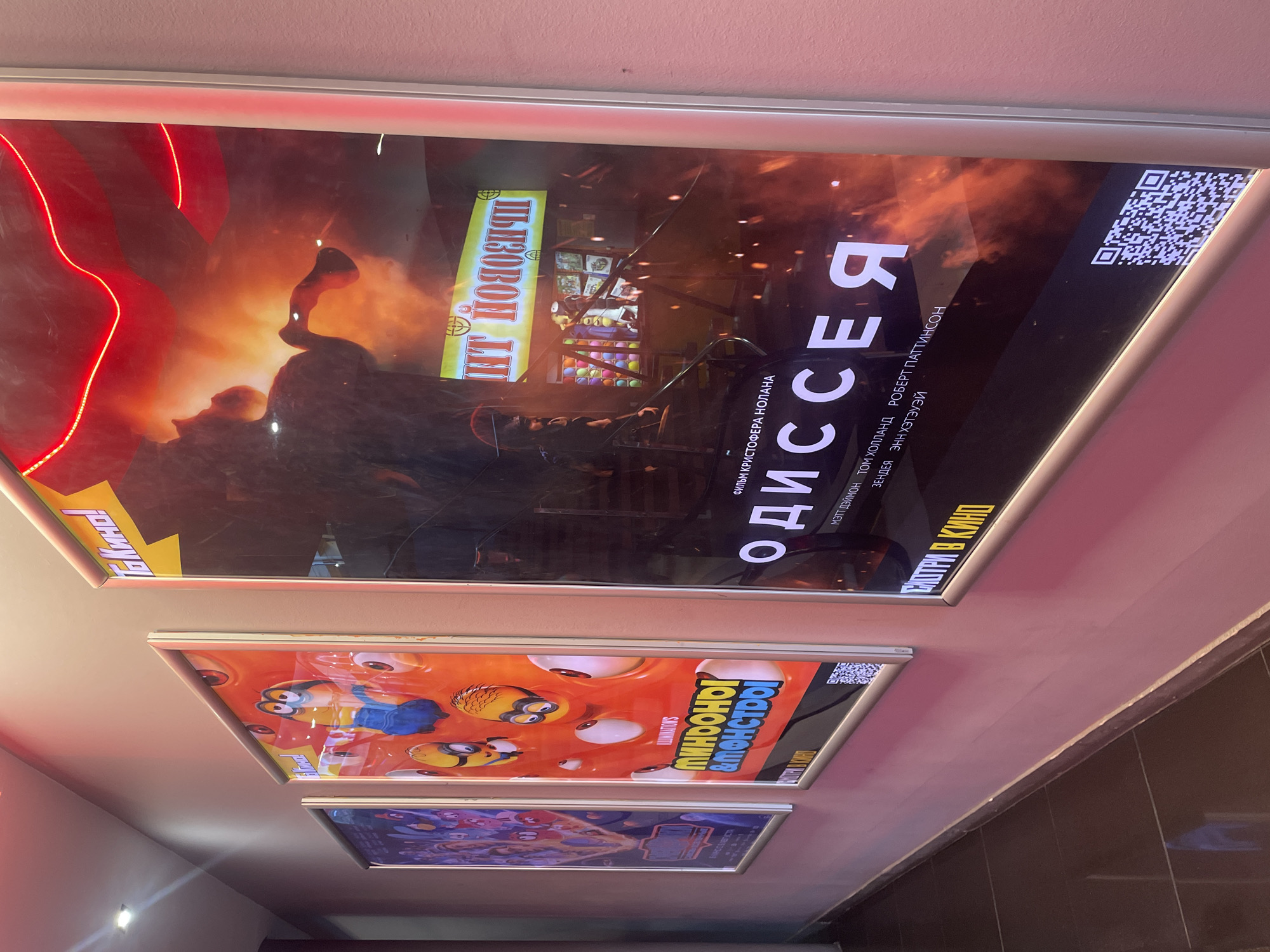 The image shows a movie theater hallway with three movie posters displayed on a pink wall. The posters are illuminated from behind.
The poster on the far left is partially visible, showing cartoon characters.
The middle poster features minions from the Despicable Me franchise, with the title "Minions & Monsters" in Cyrillic.
The poster on the right is for the movie "Oppenheimer," with its title prominently displayed in large white Cyrillic letters. Below the title are the names of actors in Cyrillic: "TOM HOLLAND, ROBERT PATTINSON." A QR code is visible at the bottom of this poster.
A red neon light strip is visible along the top of the right-hand poster. The floor is tiled in dark brown. The overall environment suggests a public indoor space, likely a cinema foyer. The city of Vaganovo, Russia, is not discernible from the visual information presented in the image.