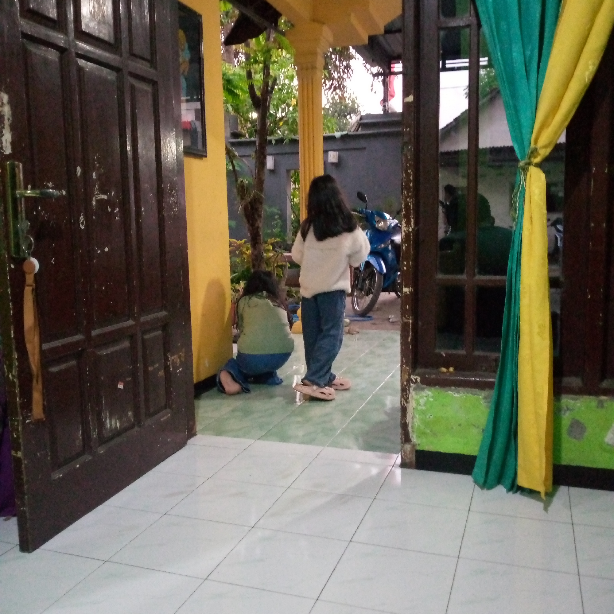 The image depicts an outdoor scene in front of a building in Sumberwono, Indonesia. Two young girls are present. One girl, wearing a white sweater and blue pants, stands facing away from the camera, looking towards a blue motorcycle parked nearby. The other girl, wearing a green shirt and blue pants, is crouched down on the tiled floor, also facing away from the camera. The setting appears to be a residential or communal area, with a brown wooden door to the left, a yellow wall, and a view of trees and structures in the background. Green and yellow fabric hangs near a window to the right.