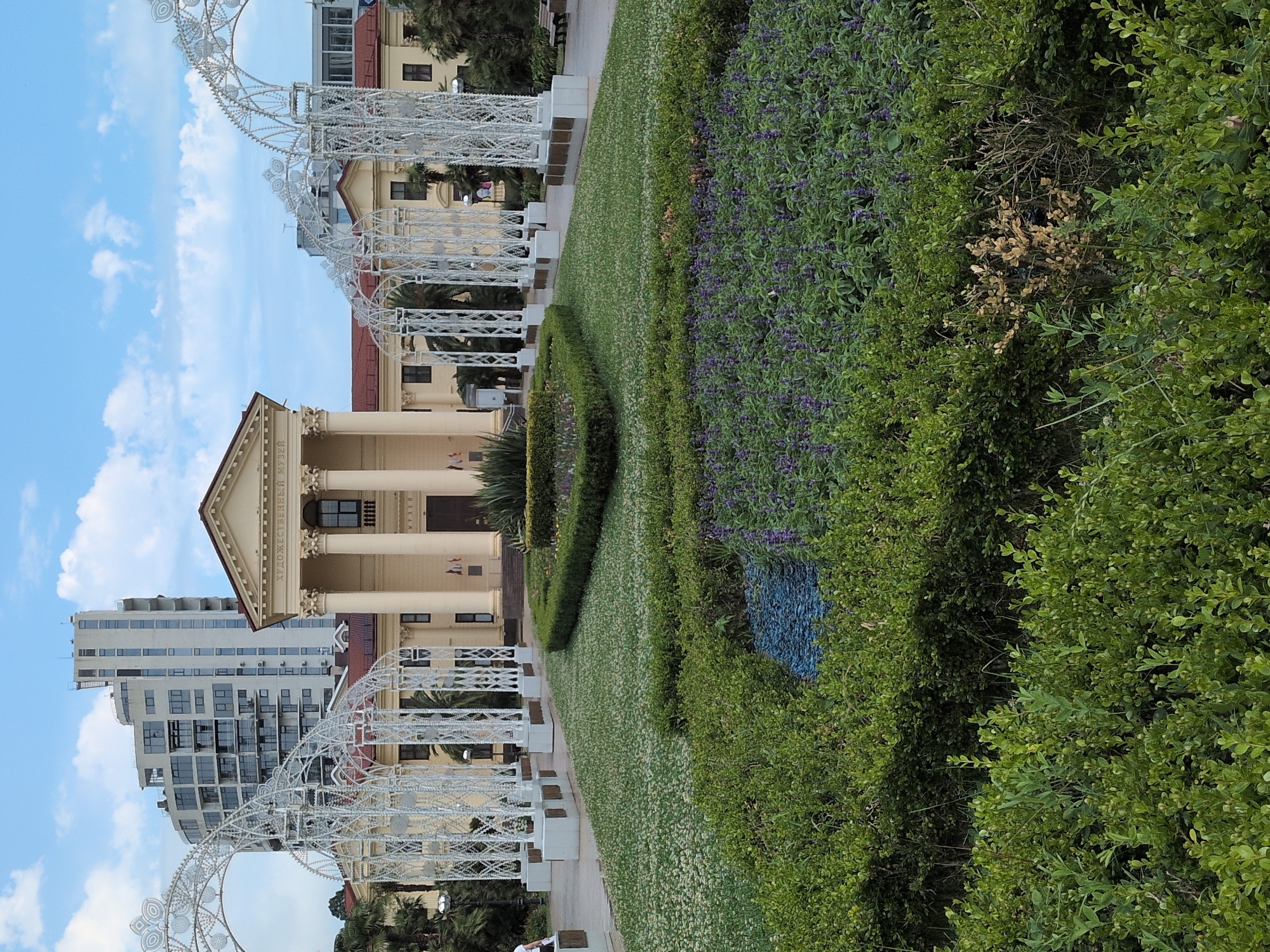 This media file depicts a vibrant outdoor scene, likely in Sochi, Russia, bathed in daylight under a partly cloudy sky. The central focus is a beautifully maintained garden with a striking architectural element. A long, low hedge frames a section of manicured lawn covered in small white flowers, which then gives way to a dense bed of purple flowering plants. To the right, a wall of lush green bushes provides a rich backdrop.
In the background, a grand, light-colored building with classical columns and a pediment stands prominently. To its left, a modern, multi-story building with a facade of numerous windows adds a contrasting architectural style. White, ornate metal structures, resembling decorative arches or gazebos, are positioned in the foreground and mid-ground, adding a touch of elegance to the landscape.
The scene suggests a public space, possibly a park or museum grounds, designed for visitors to enjoy. The presence of benches along the pathway indicates a place for relaxation and observation. The overall atmosphere is serene and picturesque, highlighting the beauty of the meticulously curated garden and the harmonious blend of historical and contemporary architecture. The time of day appears to be daytime, with the sky showing patches of blue and scattered clouds, suggesting pleasant weather. Visible text on the background building is in Russian and reads "ХУДОЖЕСТВЕННЫЙ МУЗЕЙ" which translates to "ART MUSEUM".