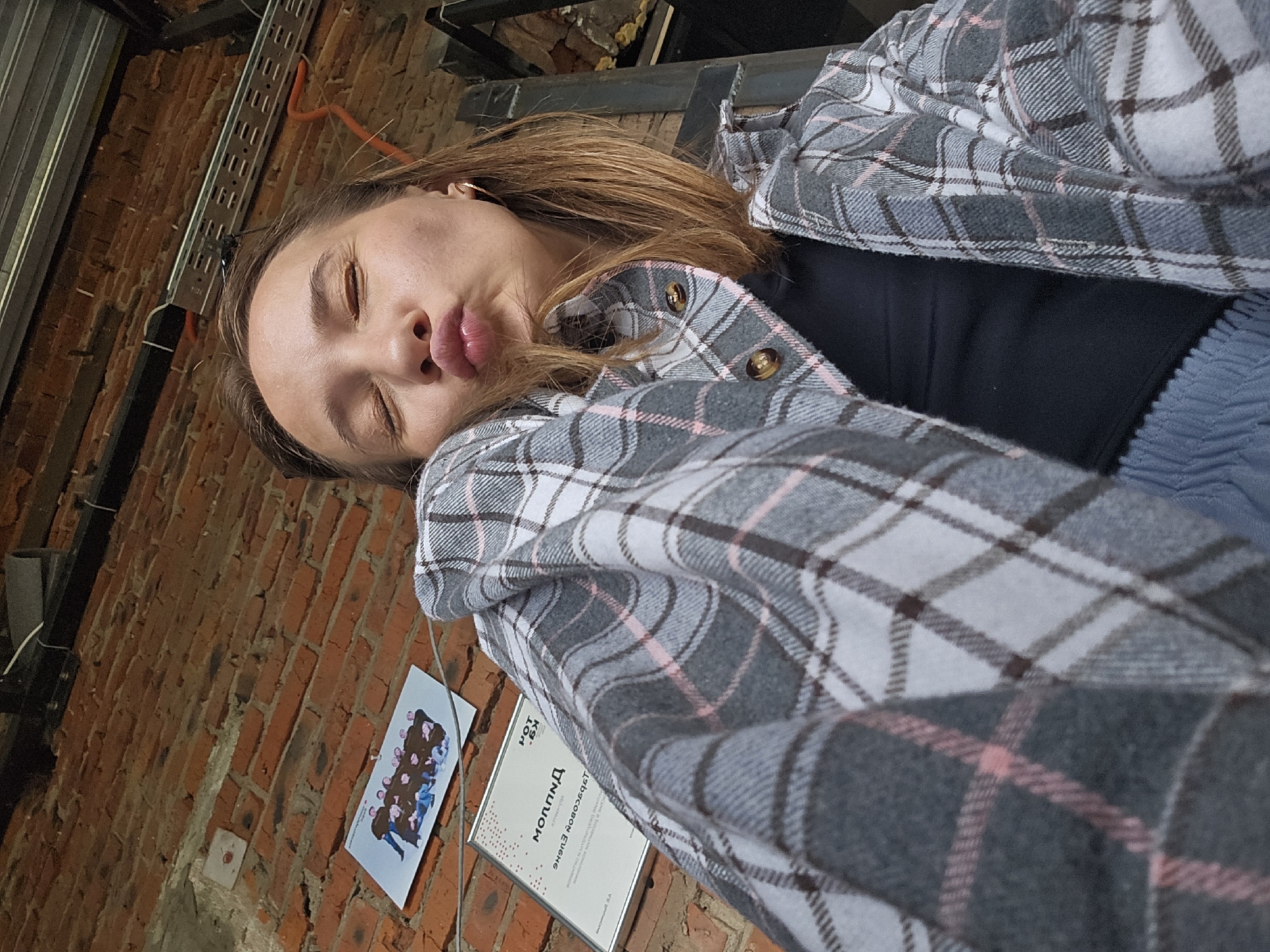 An overhead, close-up, selfie of a young woman with light brown hair is taken in front of a brick wall. The woman has her eyes closed and her lips pursed in a kissing gesture. She is wearing a grey, pink, and white plaid jacket over a black top, and blue corduroy pants. To the left of the woman, on the brick wall, are two pieces of paper. The upper paper is a photograph of a group of people wearing graduation gowns. The lower paper is a certificate or award. The brick wall is rough and textured, with visible mortar lines. Above the wall, a metal structure with wires and conduit is visible. The background is dark and out of focus, suggesting an indoor or dimly lit outdoor setting. The overall impression is casual and playful.