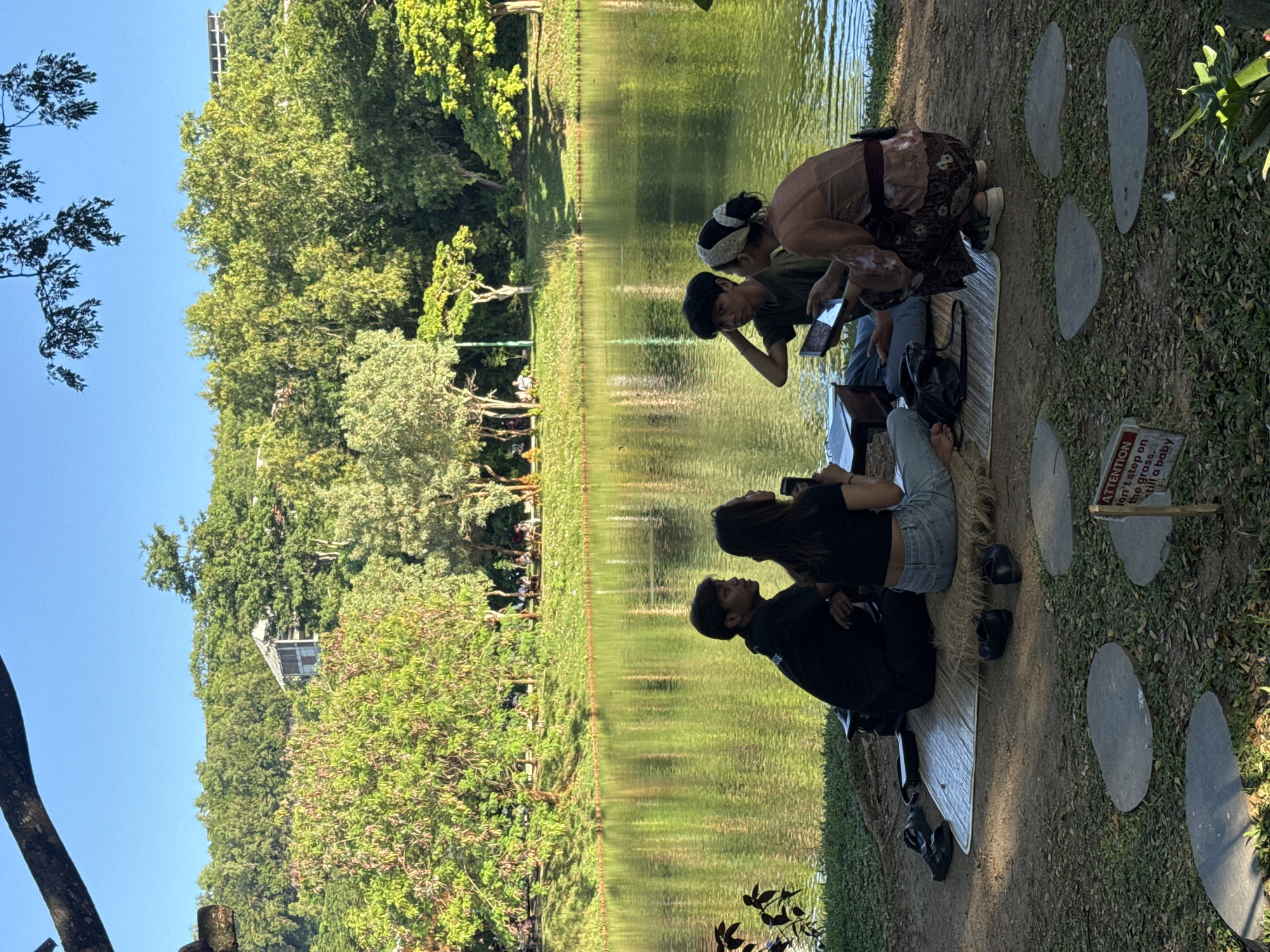 At a location in Kuta Selatan, Indonesia, a group of four people sit by a body of water. Two people are looking at a tablet device. One person is lying down, and the other is sitting up with their hand on their head. The individuals are positioned on a mat. In the background, there is lush vegetation and trees. The environment appears to be outdoors on a sunny day. There are stone pathways nearby with a small sign.